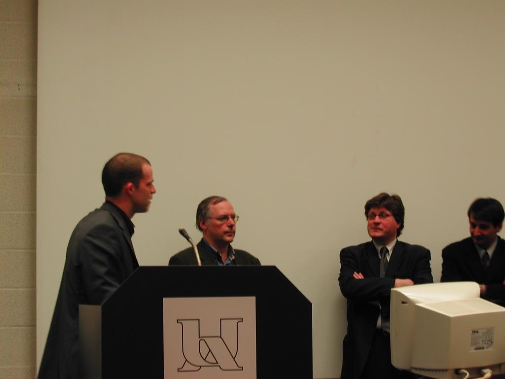 A man speaks at a podium during a PhD defense, while three others listen in the background.
