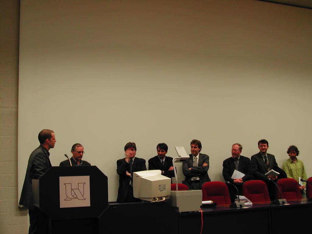 A person stands at a podium speaking to a panel of people during a PhD defense presentation.