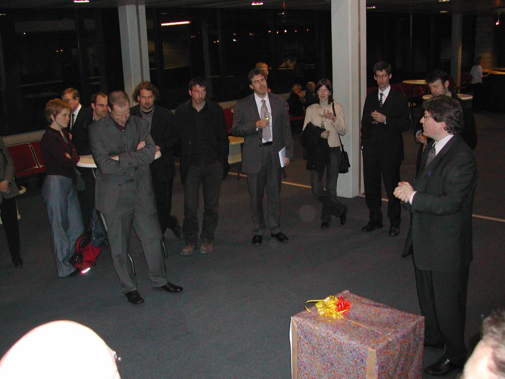 A man in a suit speaks to a group of people at a PhD defense gathering, with a wrapped gift nearby.