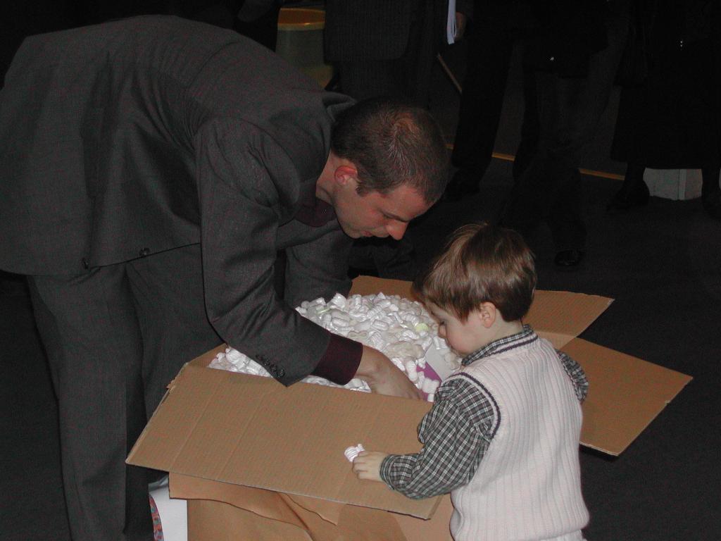A man in a suit and a young child look inside a large open box filled with packing material.