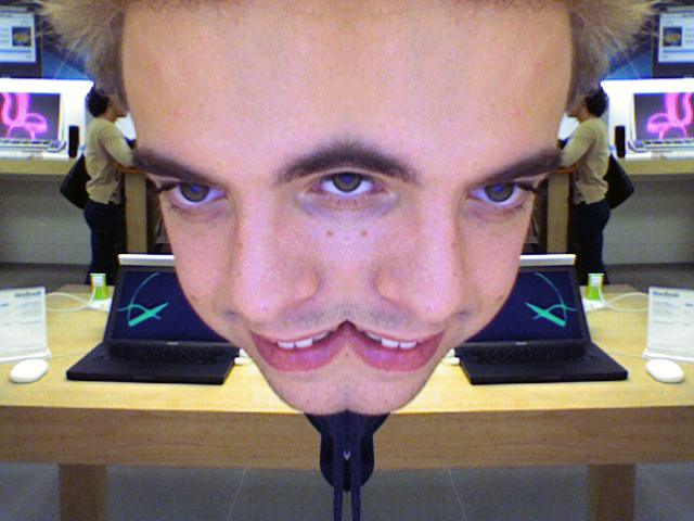 A person's face is distorted by a photo booth effect, creating a mirrored illusion with one large eye and mouth.