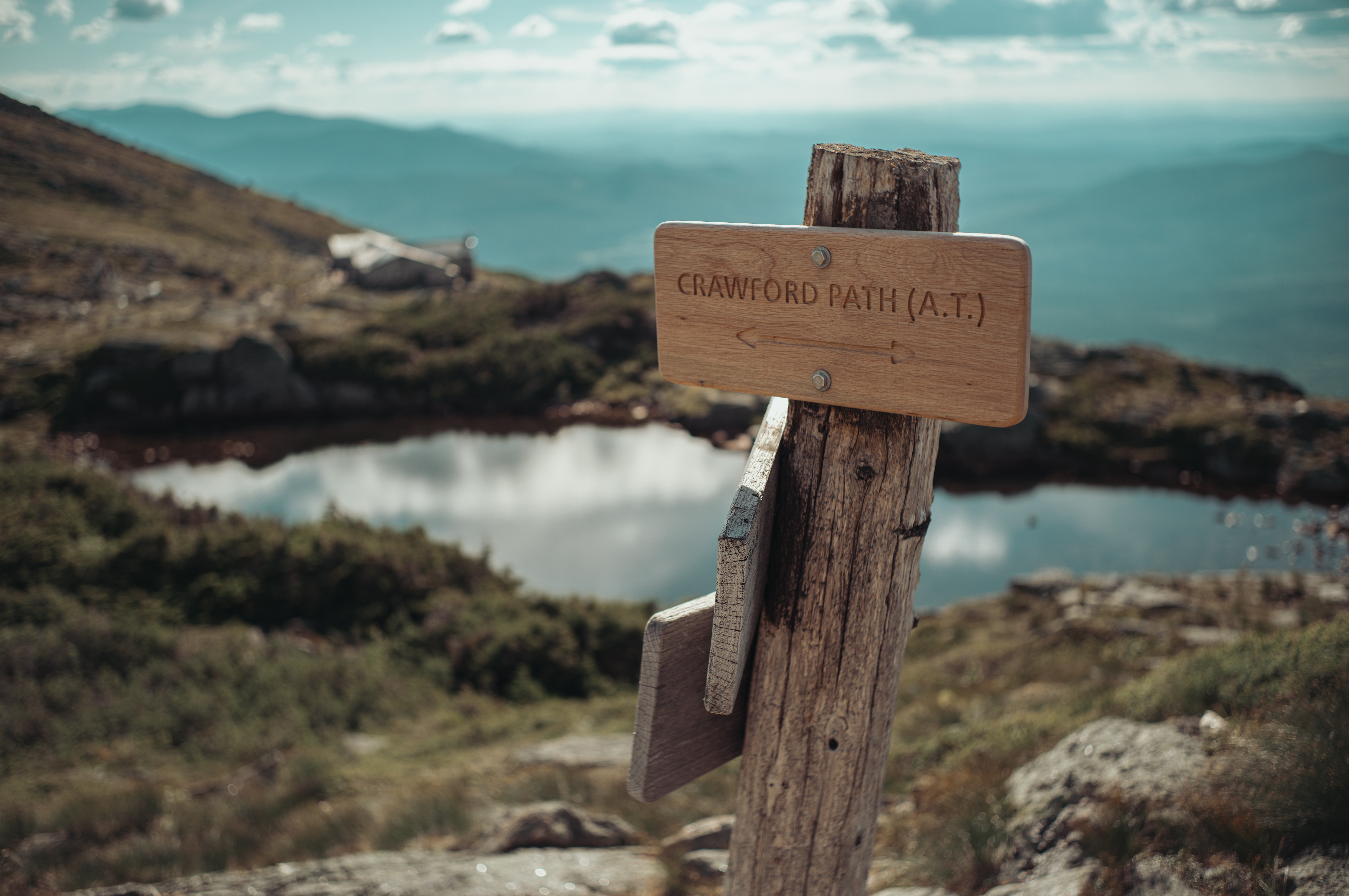 Wooden trail sign reading "Crawford Path (AT)" atop a mountain, with an alpine lake and hazy ridgelines in the background.