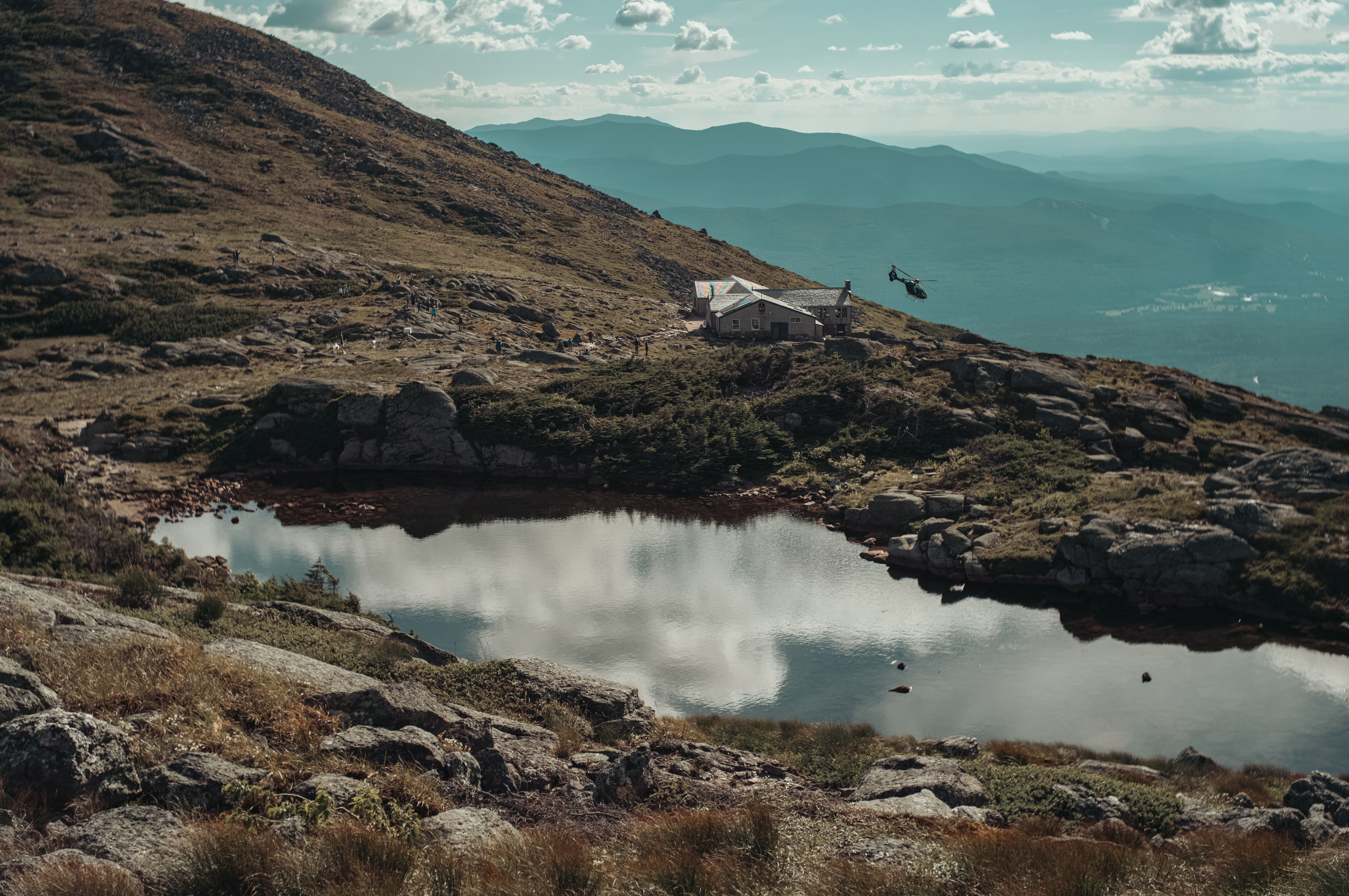 A helicopter flies near a mountain hut and alpine pond, with rocky terrain and hazy ridgelines in the background.