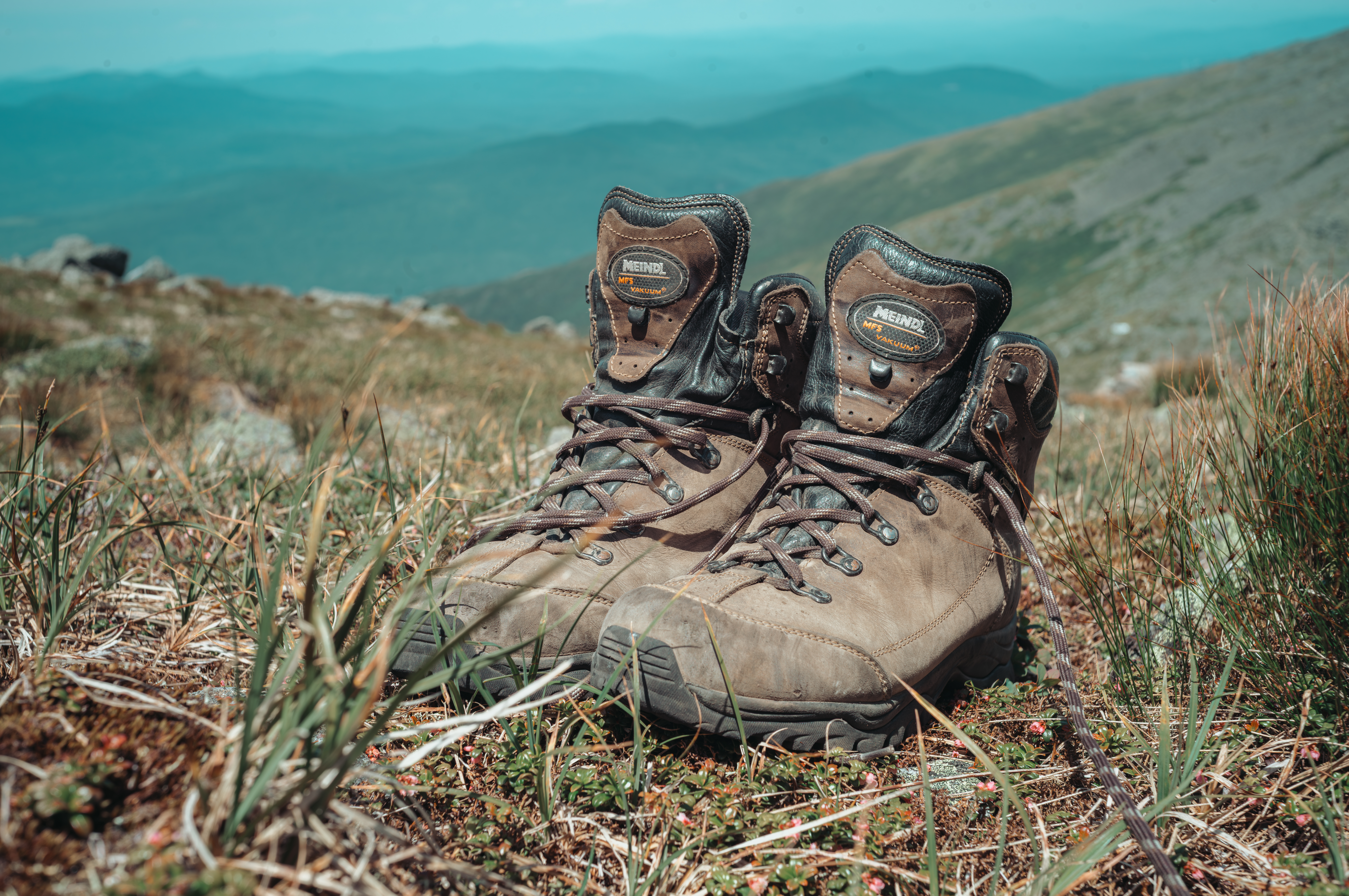 A pair of worn brown leather hiking boots rests on a grassy hillside, with mountain ranges in the distance.