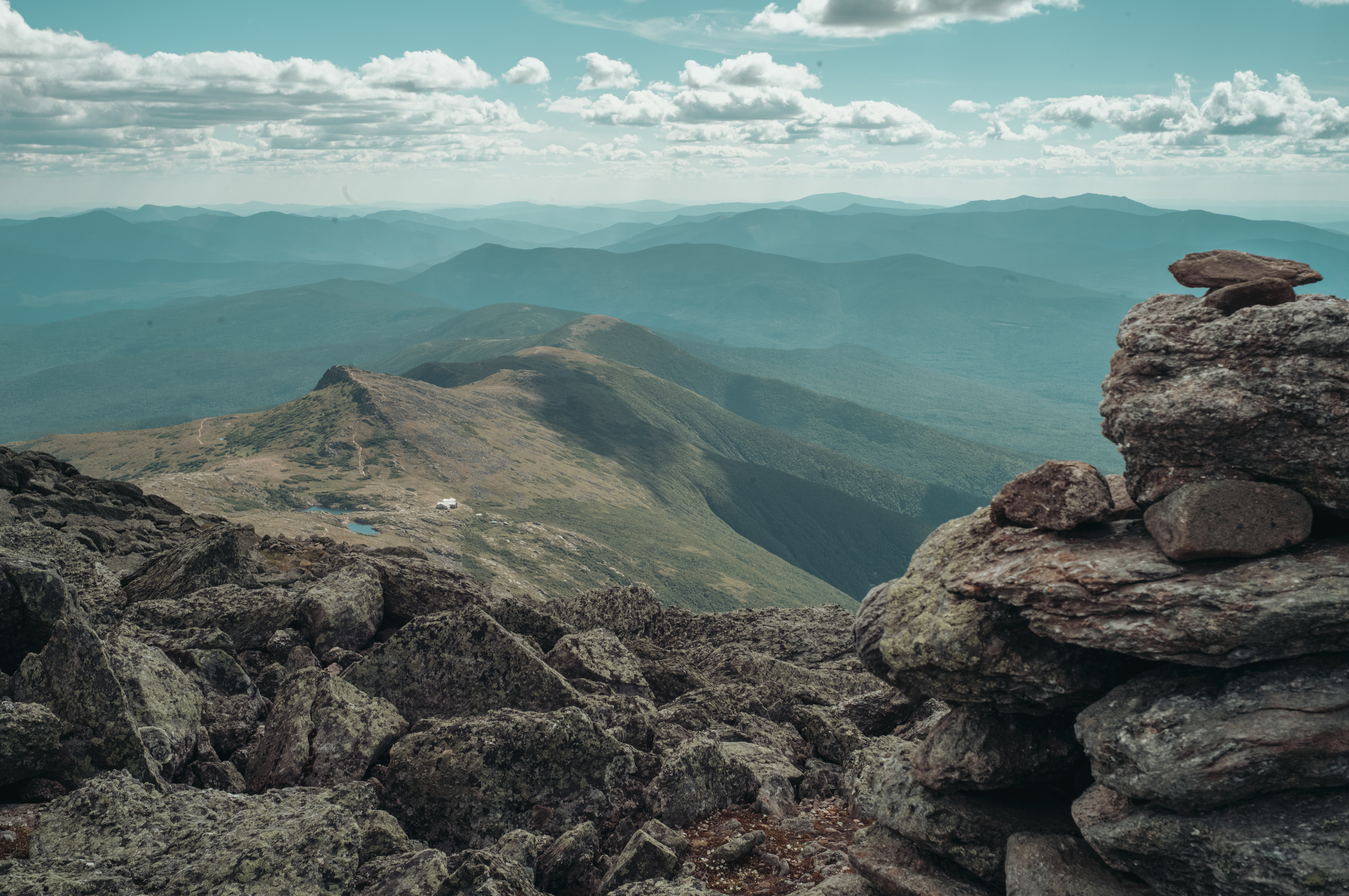 A stacked stone cairn marks a rocky mountain trail overlooking hazy ridges and a small white hut in the valley below.