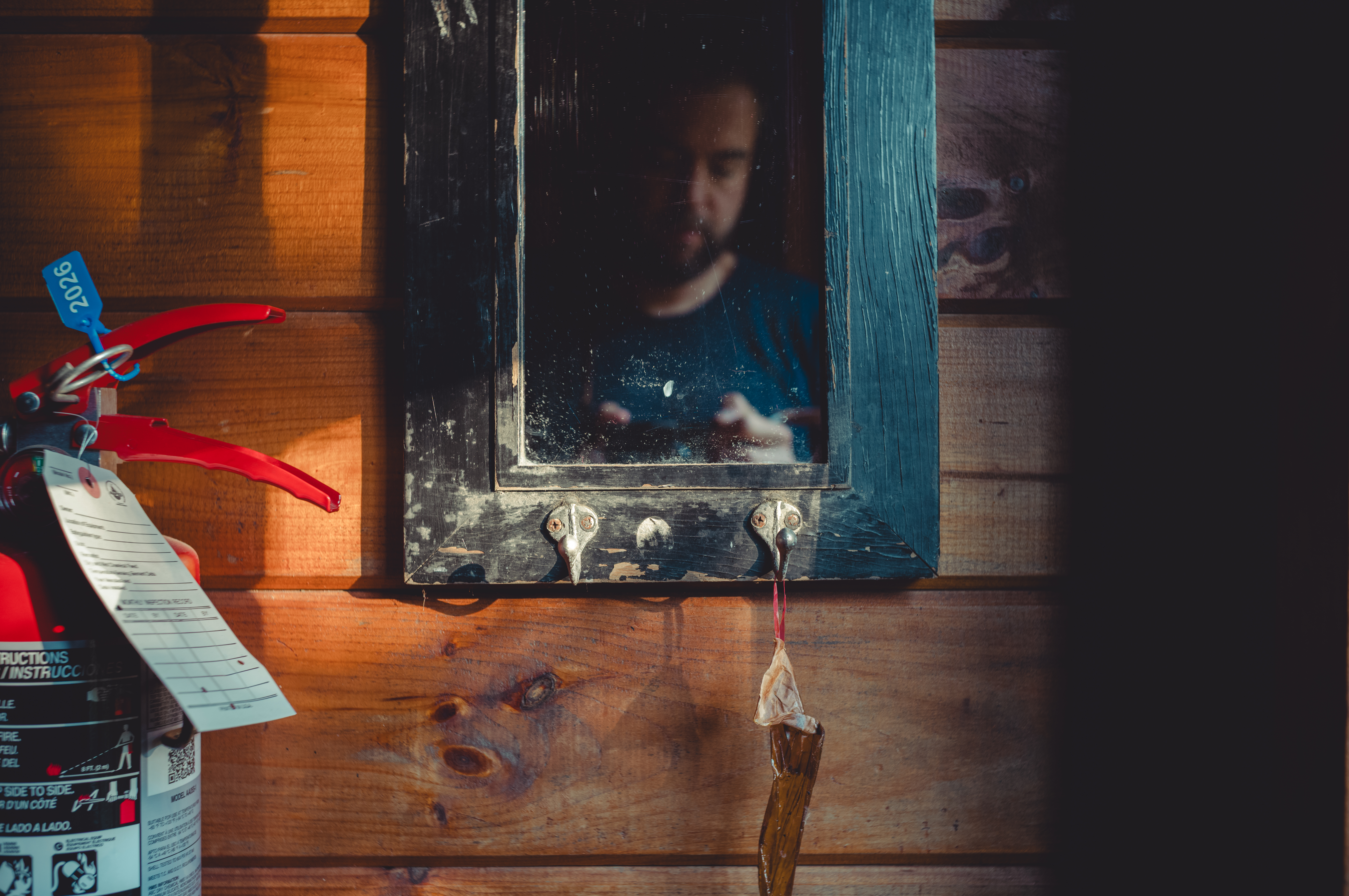 A bearded man reflected in a weathered mirror with hooks, mounted on a wooden wall beside a red fire extinguisher.