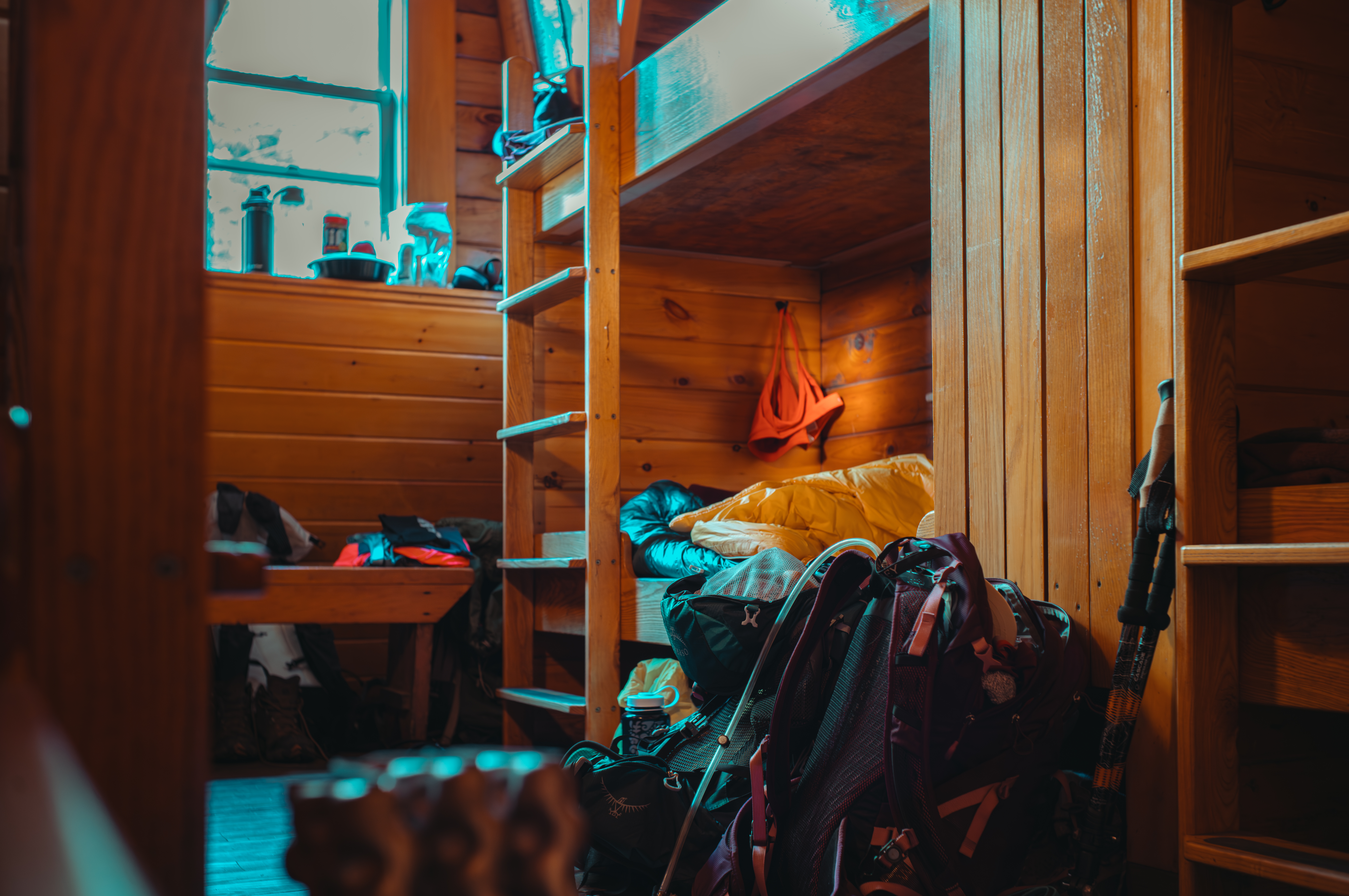 Wooden bunk beds in a cabin with backpacks, sleeping bags, and hiking gear scattered around.