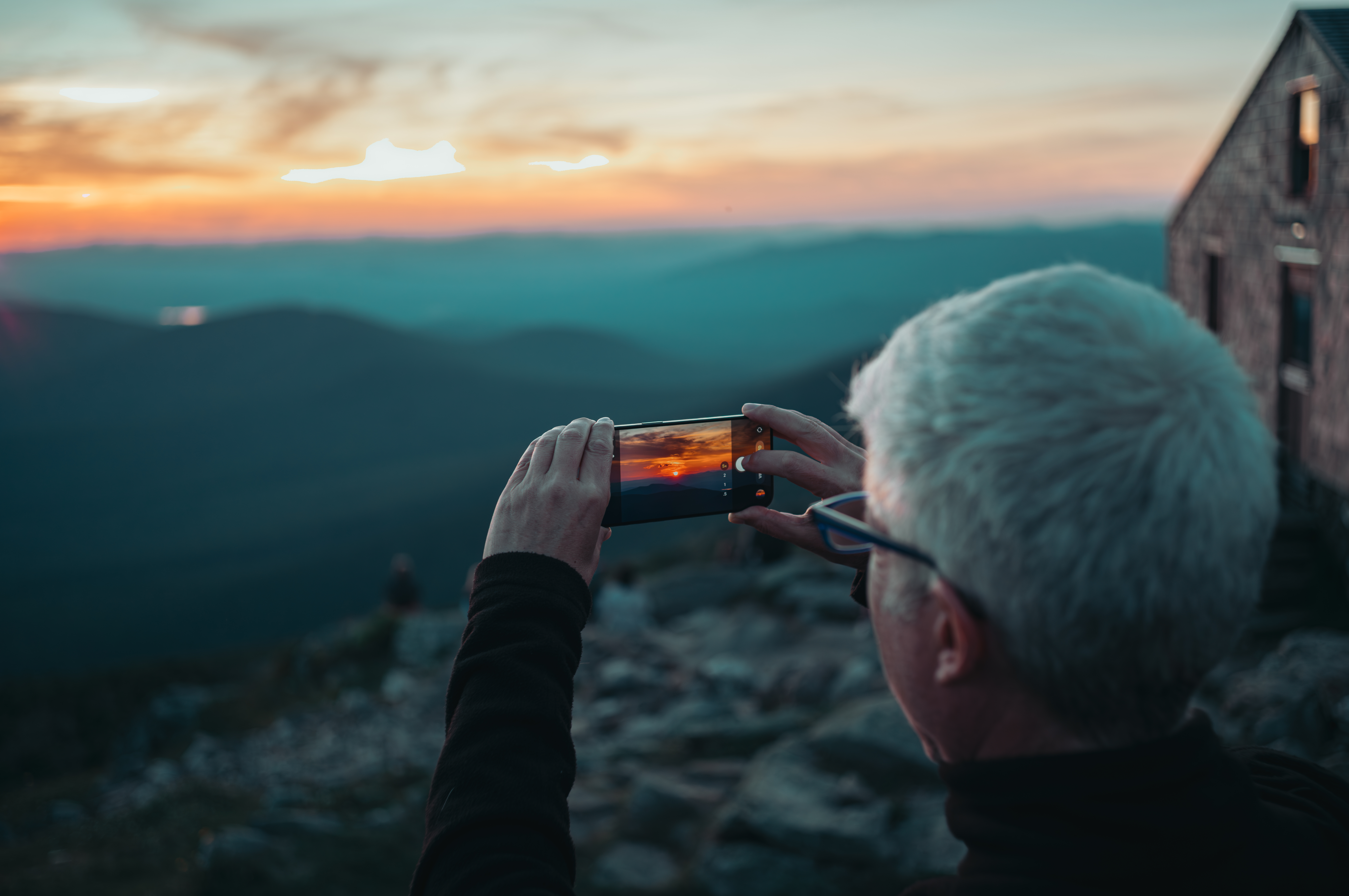 A person uses their phone to photograph a mountain sunset, with a hut visible to the right.