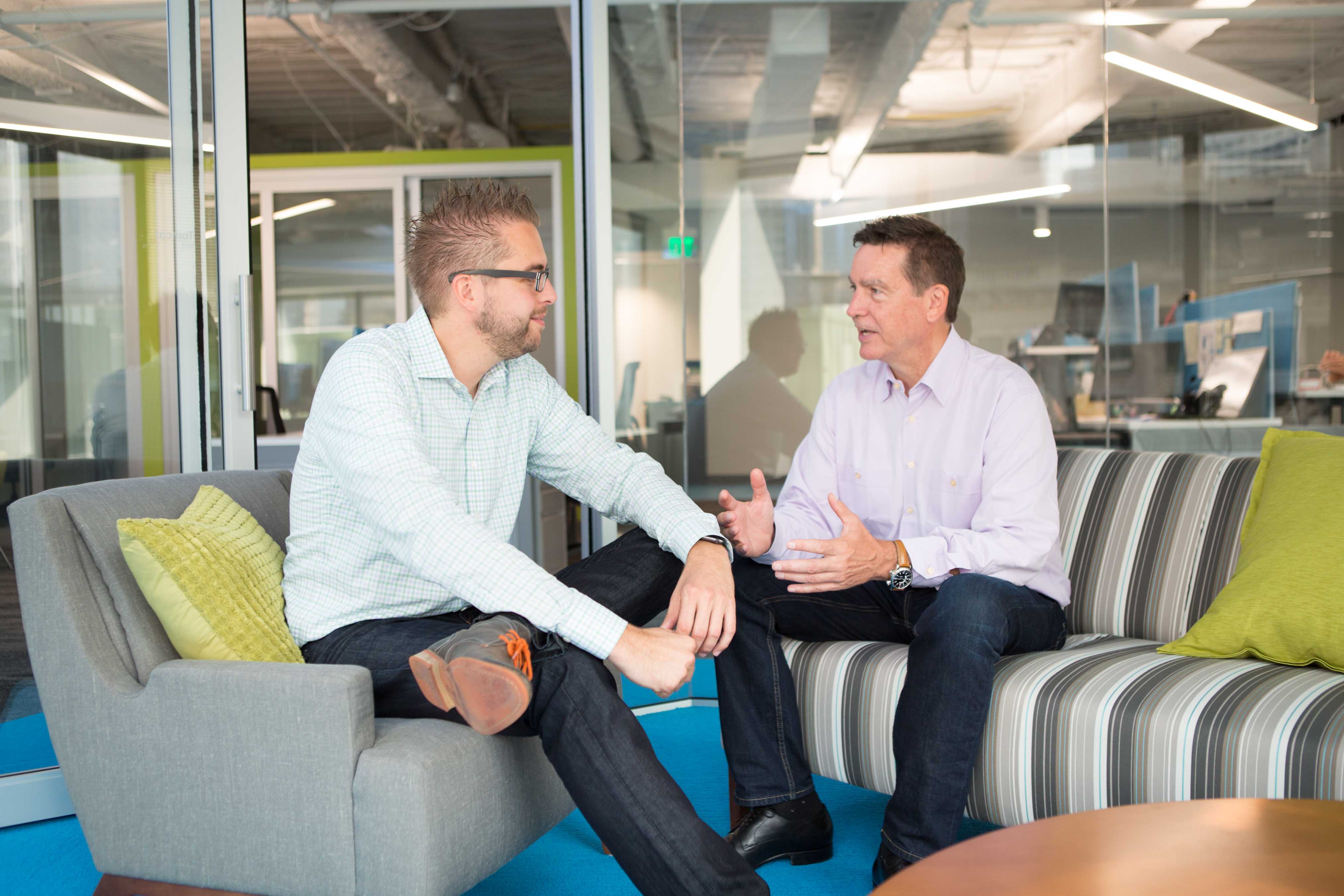 Two men sit on office lounge furniture, engaged in a conversation.