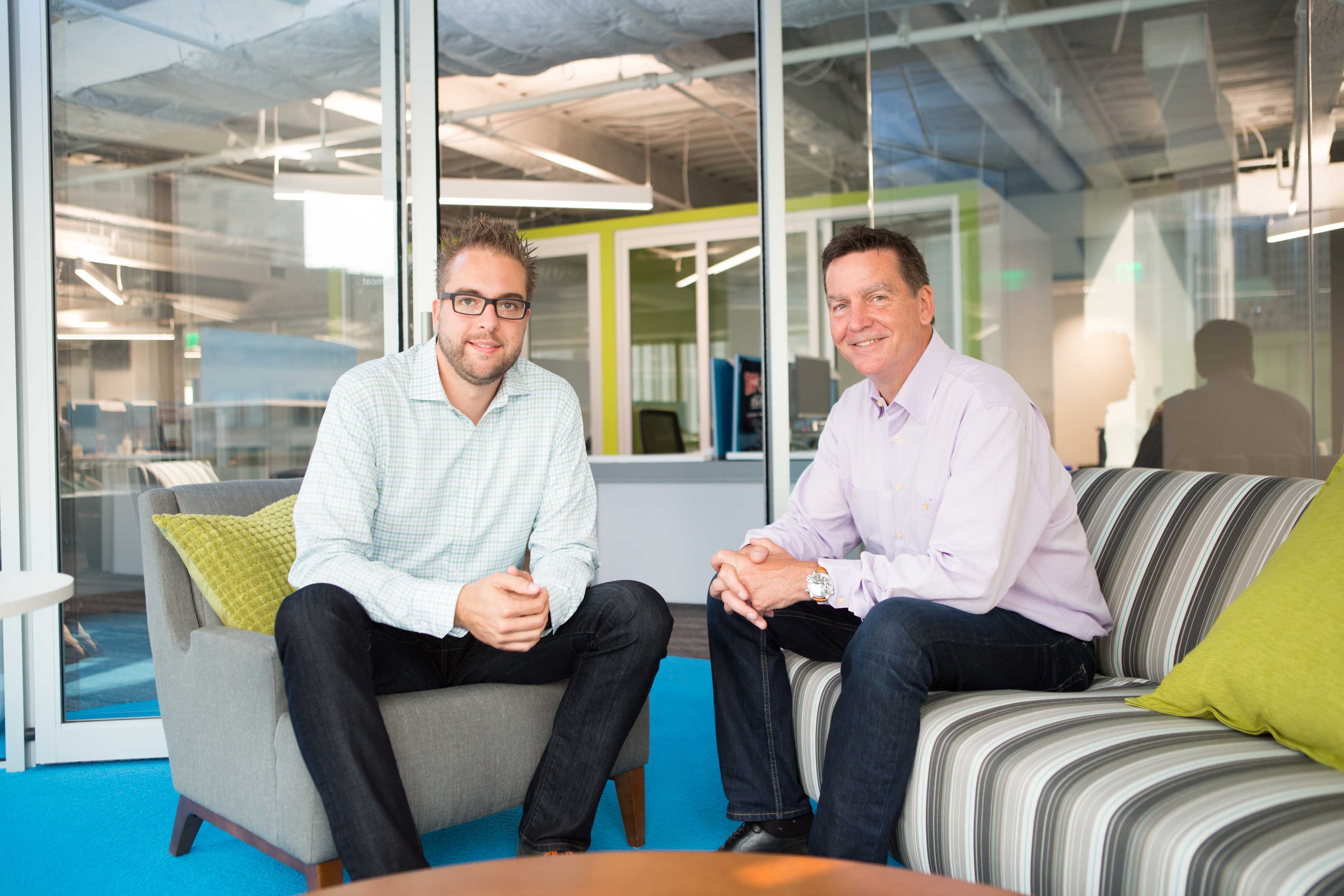 Two men sit in a modern office lounge, smiling at the camera.