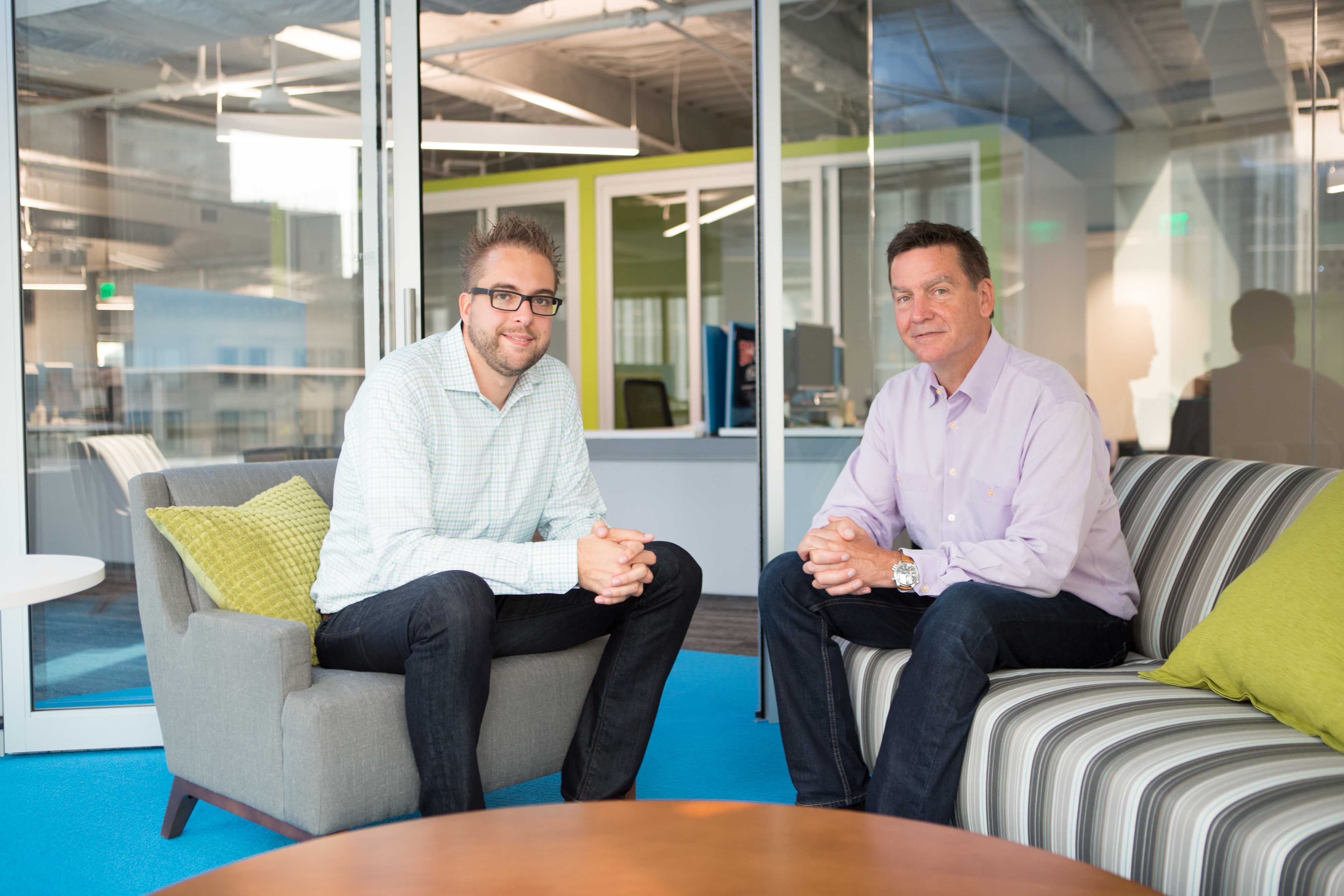 Two men sit on office lounge chairs, smiling at the camera in a modern workspace with glass walls.
