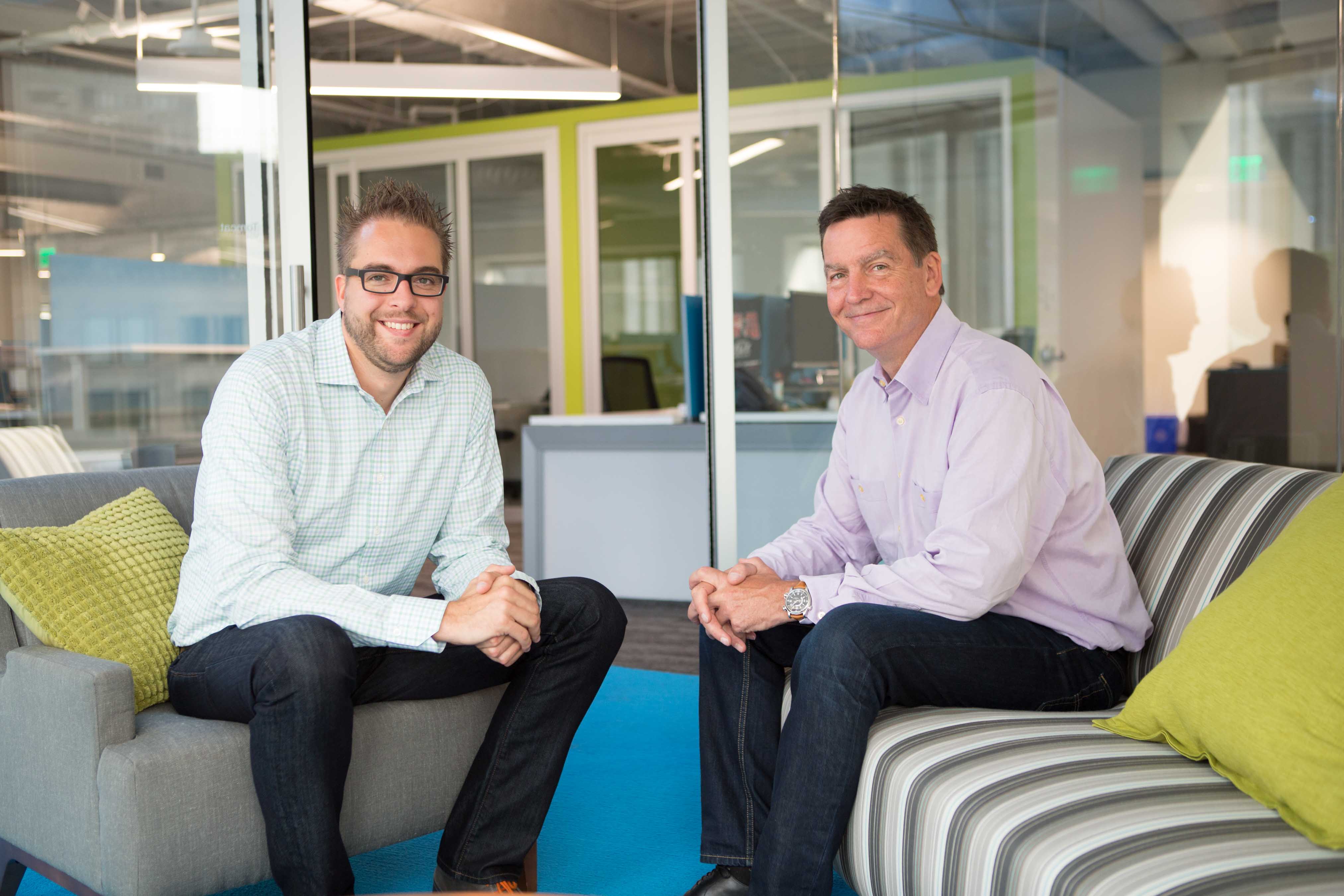 Two men sitting in a modern office lounge, smiling at the camera.