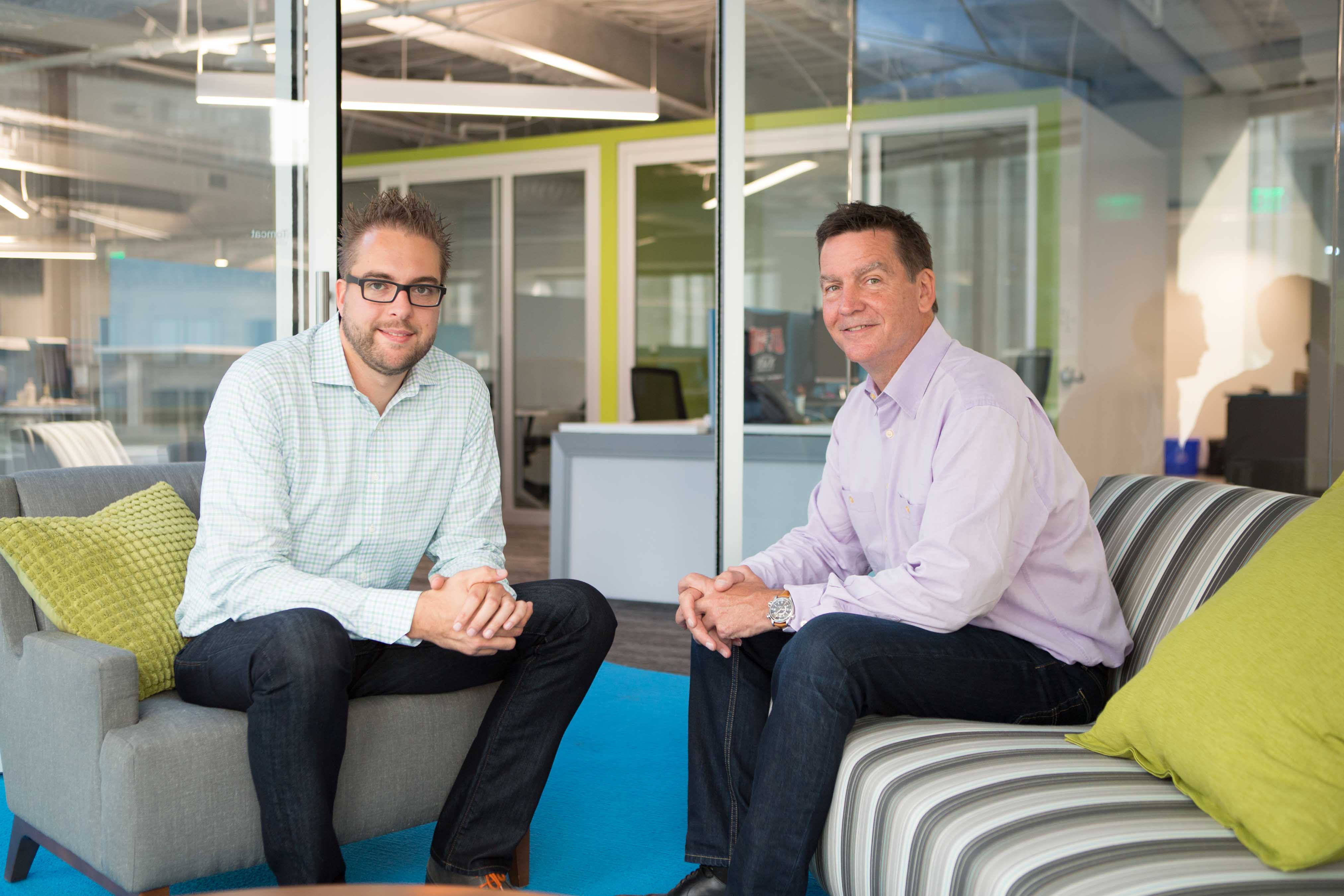 Two men sitting in a modern office lounge, smiling at the camera.