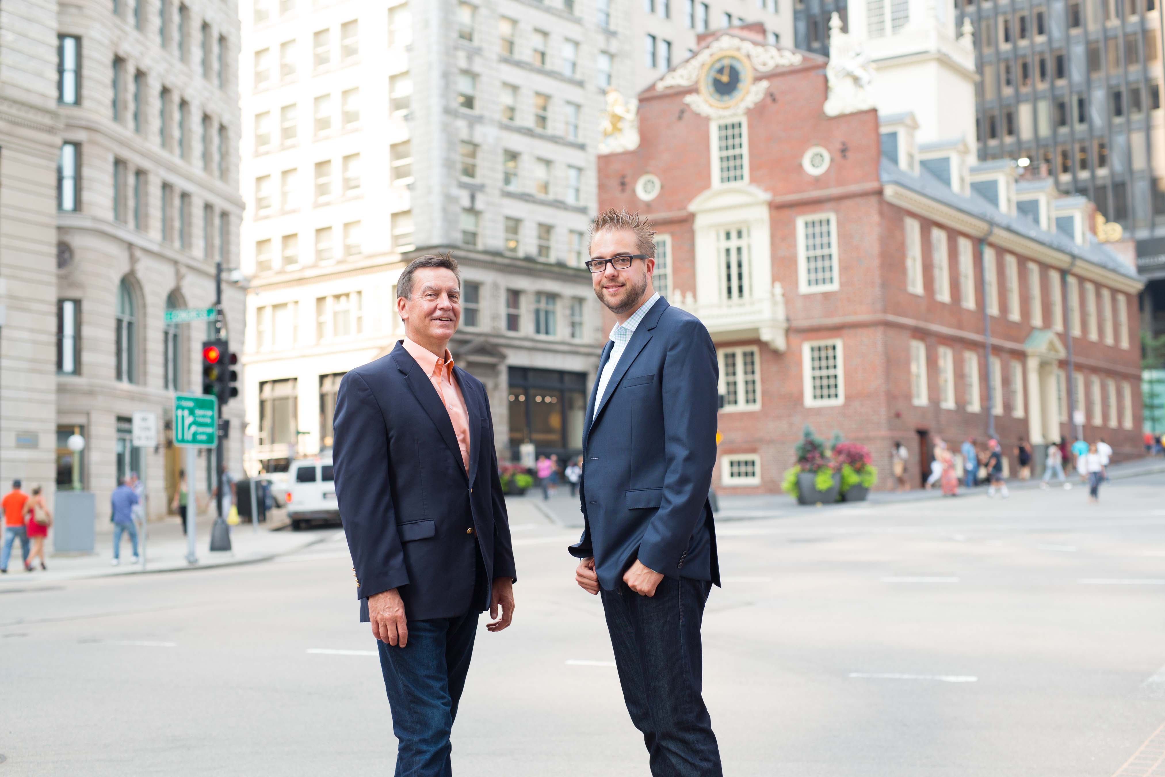 Two men in business attire stand on a city street, posing for a professional photo with historic buildings behind them.