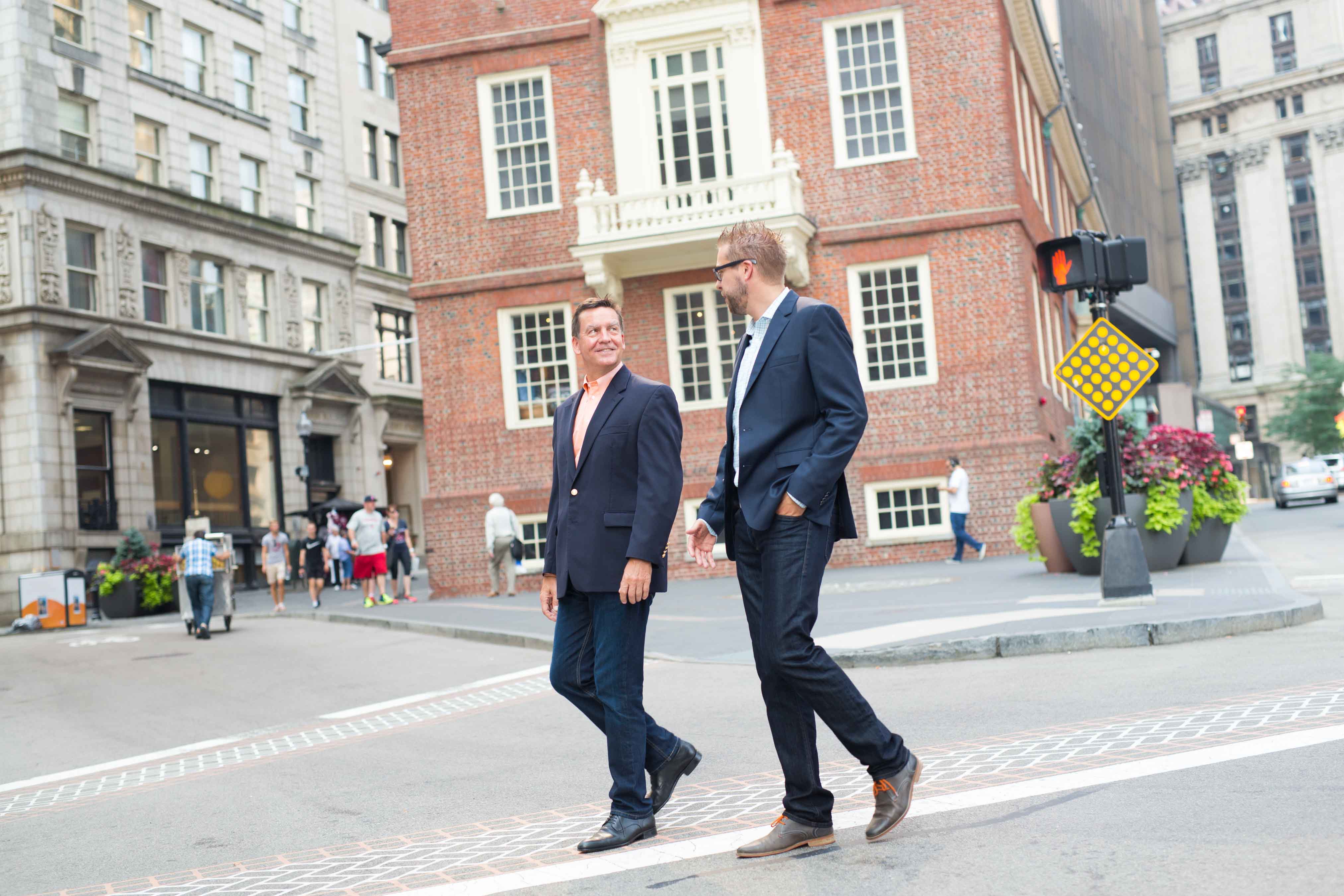 Two men in business attire walk and talk on a city street near a historic brick building.