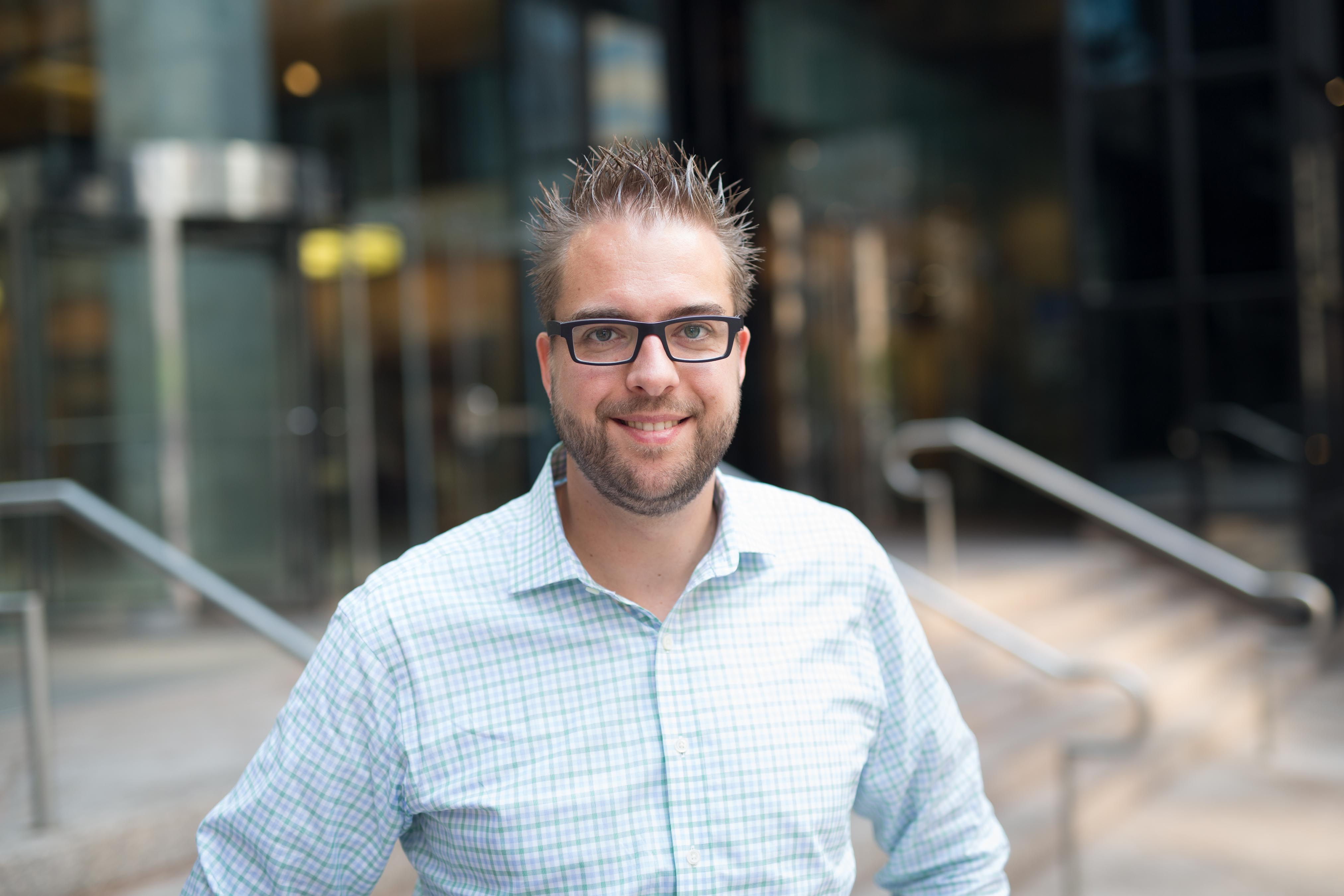 A man wearing glasses and a checkered shirt smiles while standing outside a modern office building.