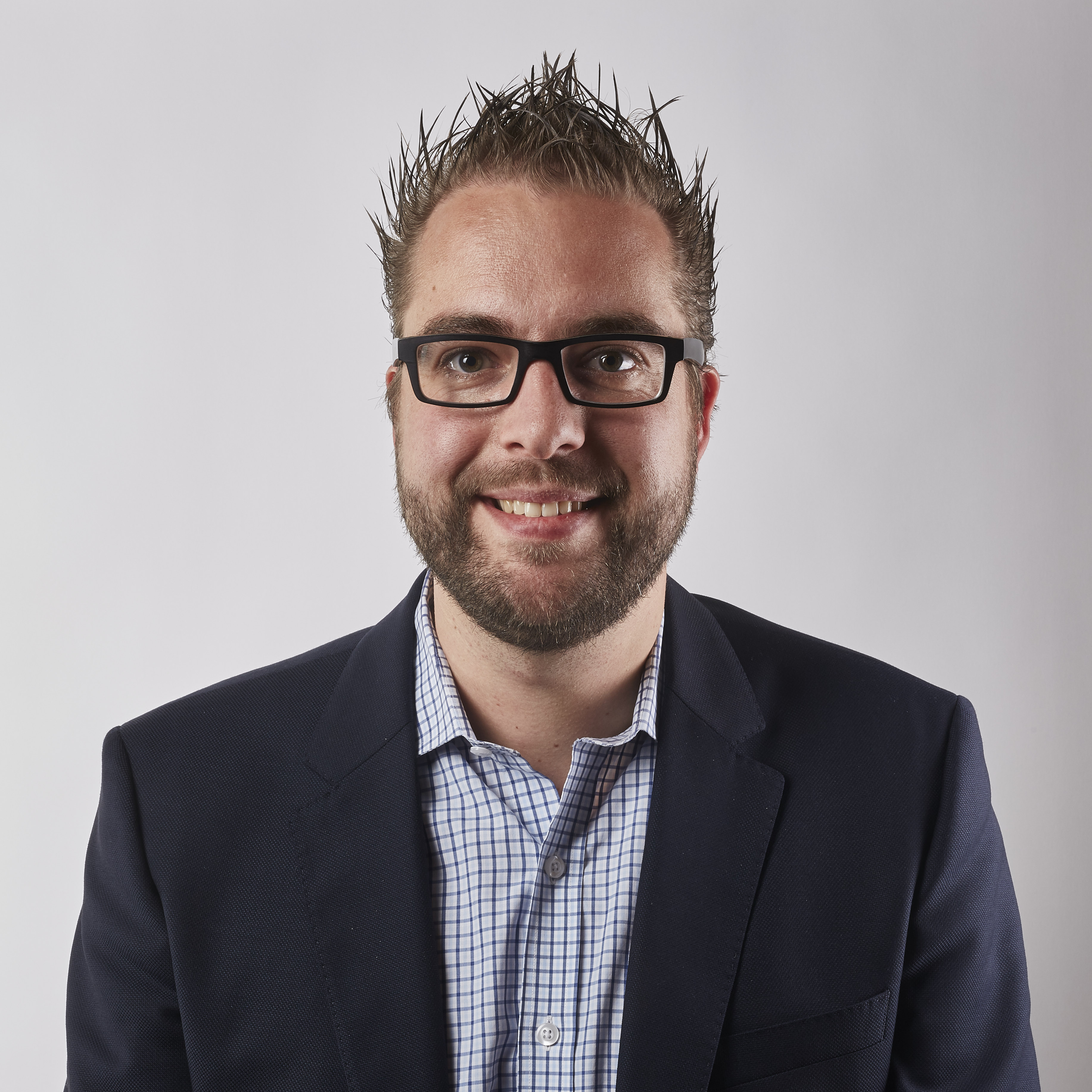 A man wearing glasses and a suit jacket smiles at the camera against a plain white background.