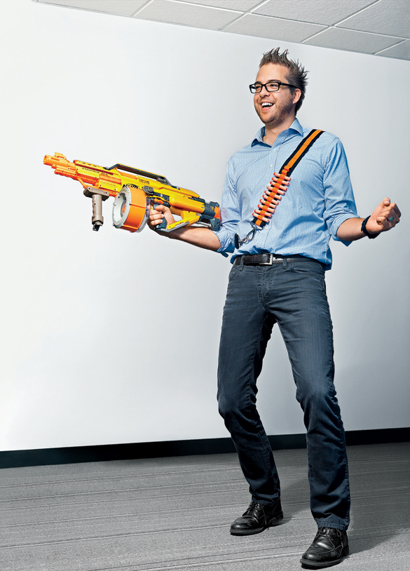 A man wearing glasses and a blue shirt holds a large Nerf gun and smiles excitedly.