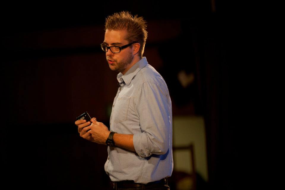 A speaker in a light blue shirt and glasses presents on stage at DrupalCon London, holding a remote control.