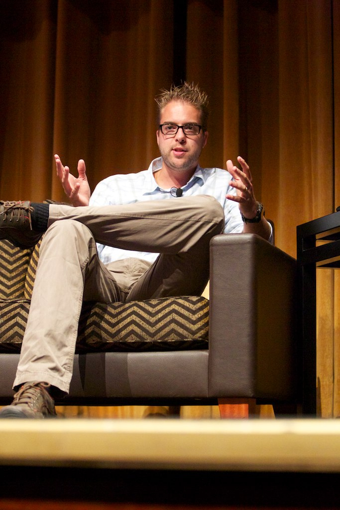 A man sits on a stage chair, gesturing with both hands while speaking at DrupalCon Munich.