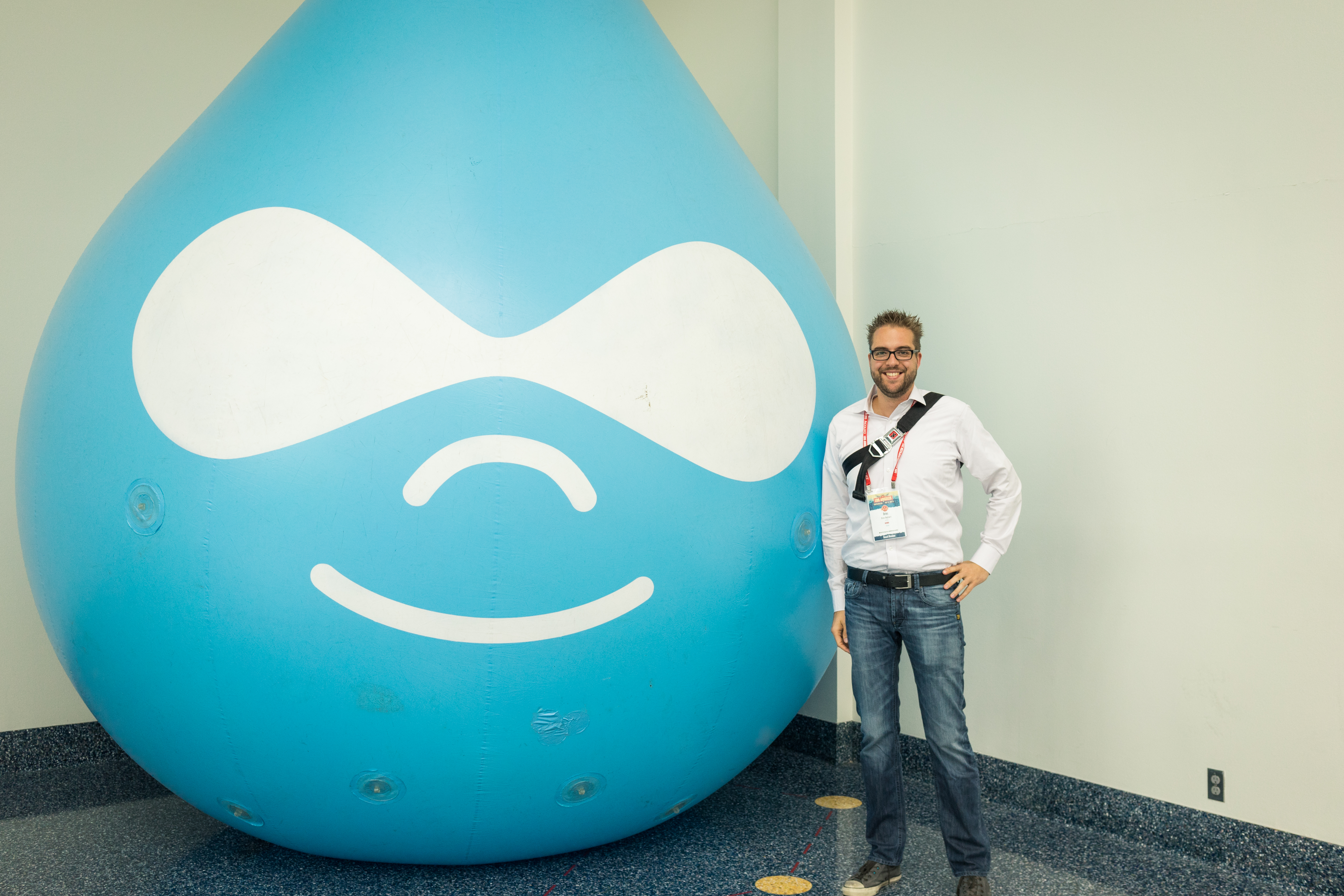 A man in a white shirt stands next to a large inflatable Druplicon mascot, smiling at the camera.