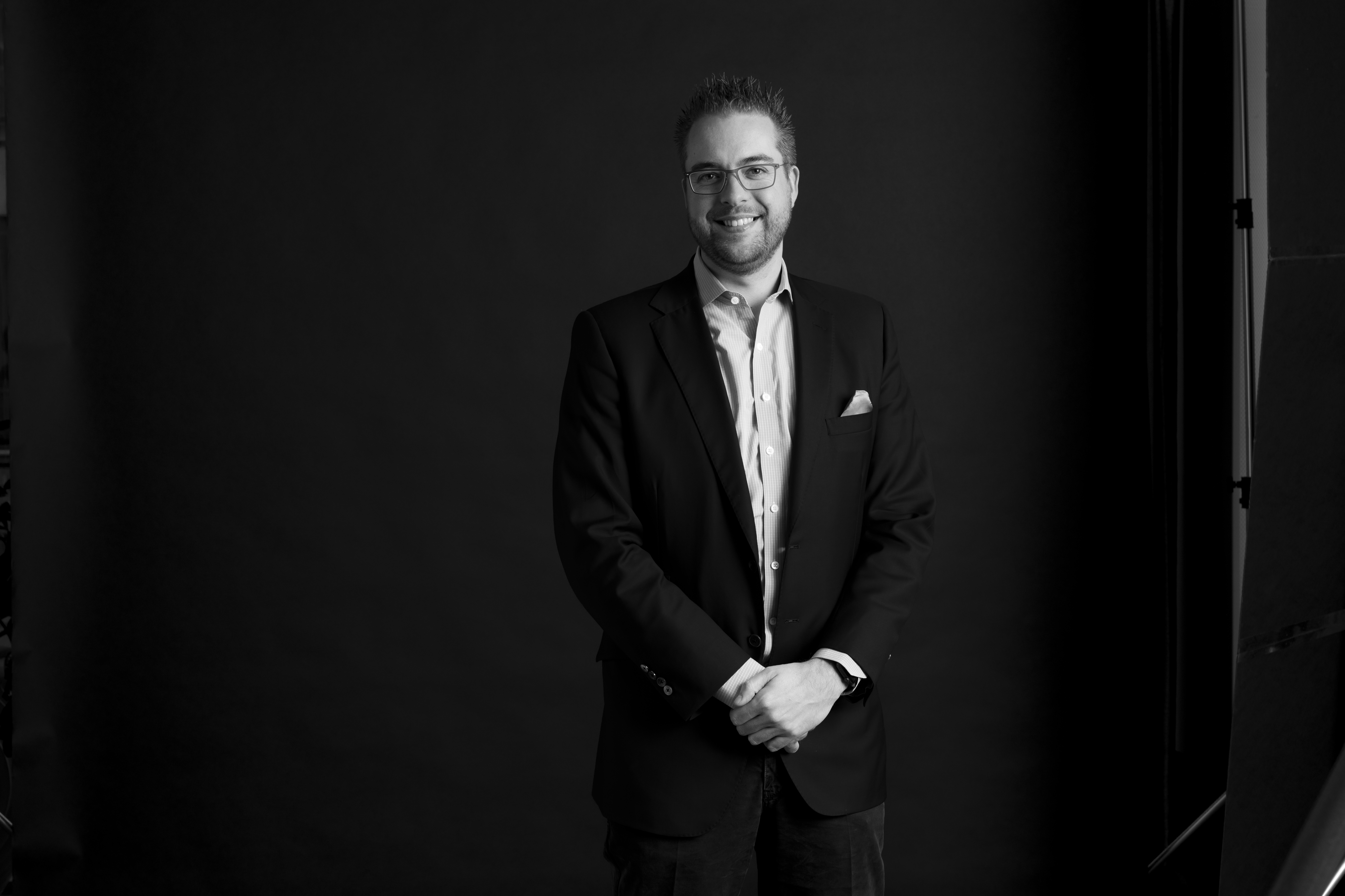 A man in a suit and glasses stands against a dark background, smiling at the camera.