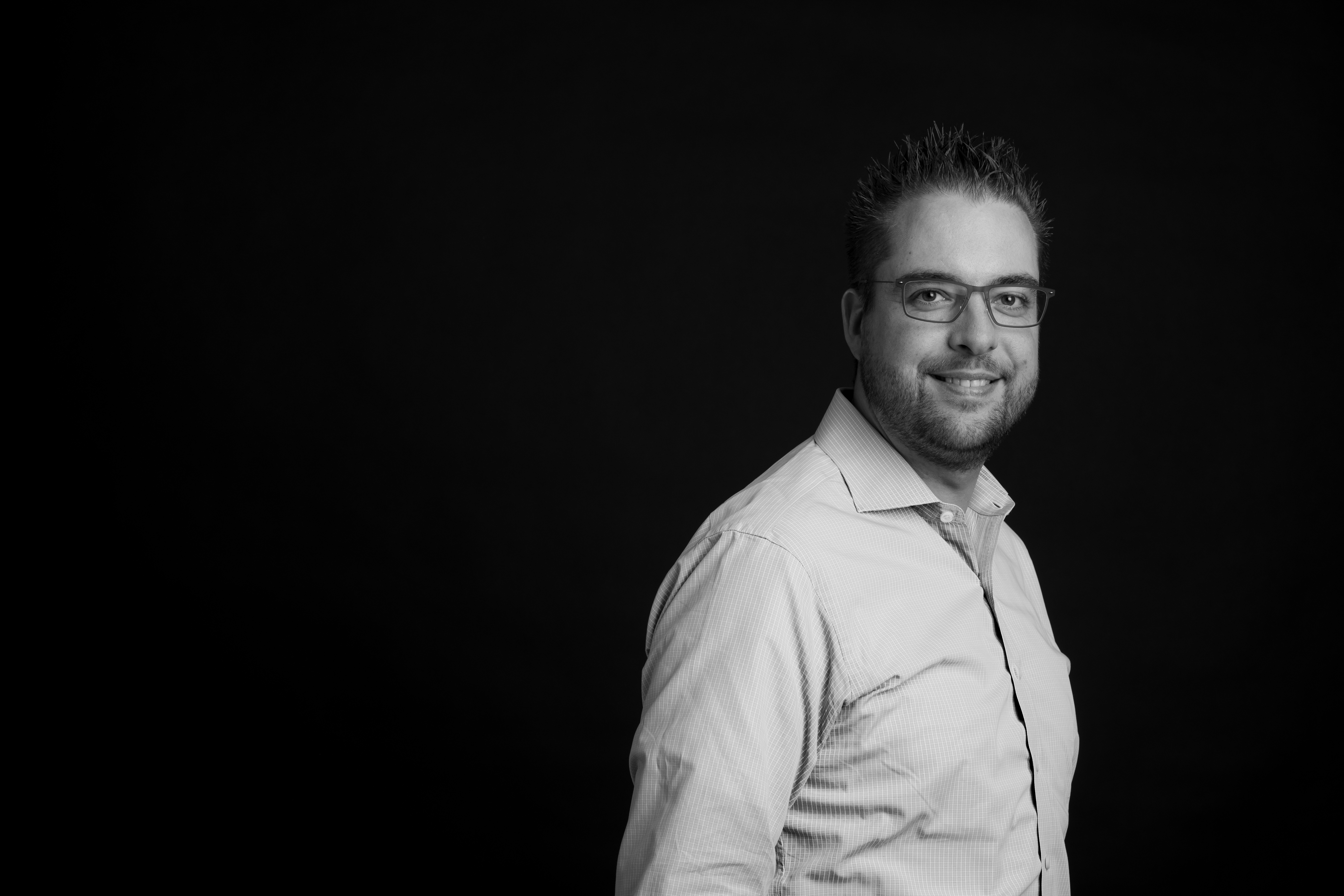A man wearing glasses and a button-up shirt smiles while posing against a dark background.