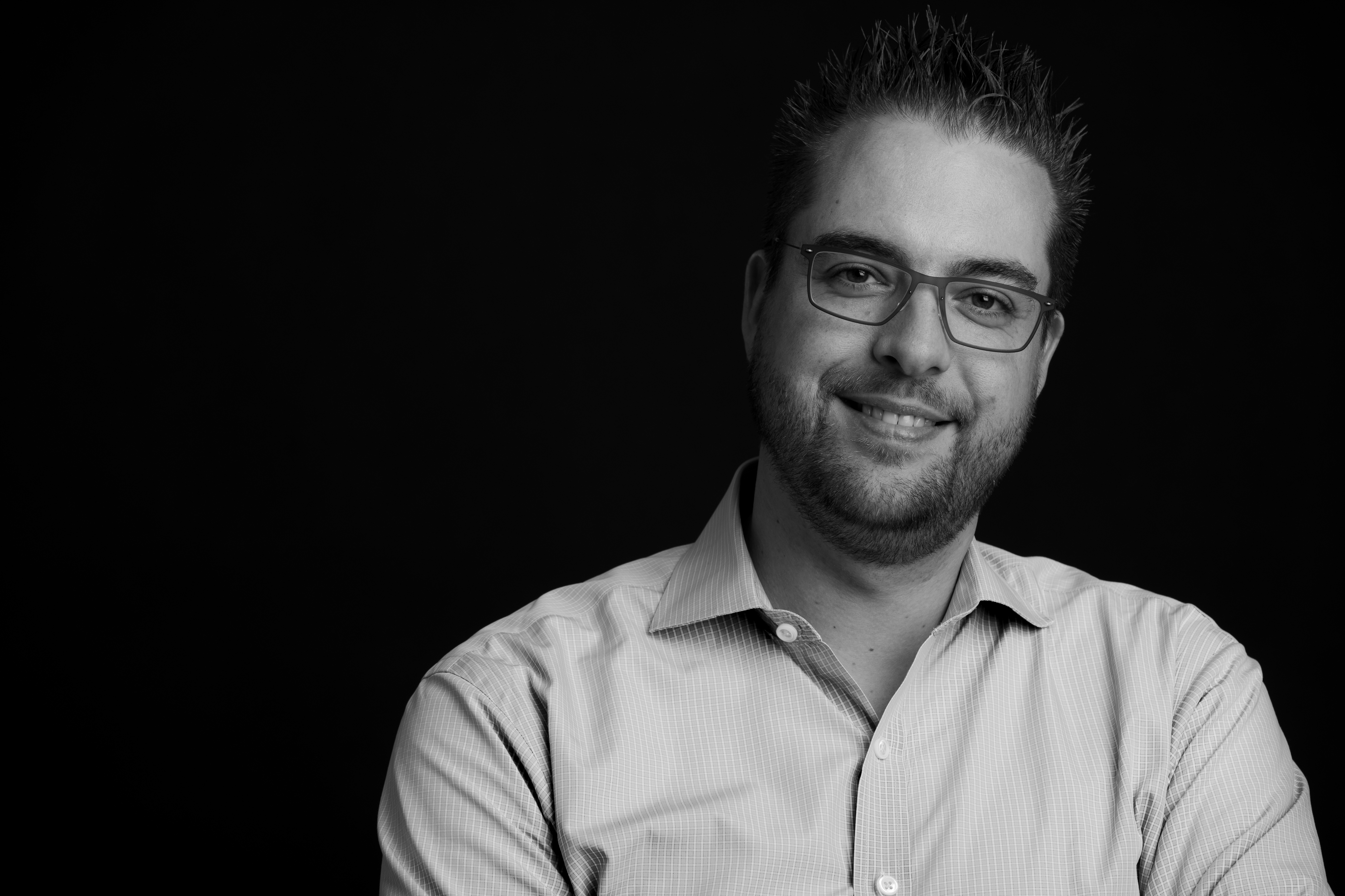 A man wearing glasses and a button-up shirt smiles at the camera against a dark background.