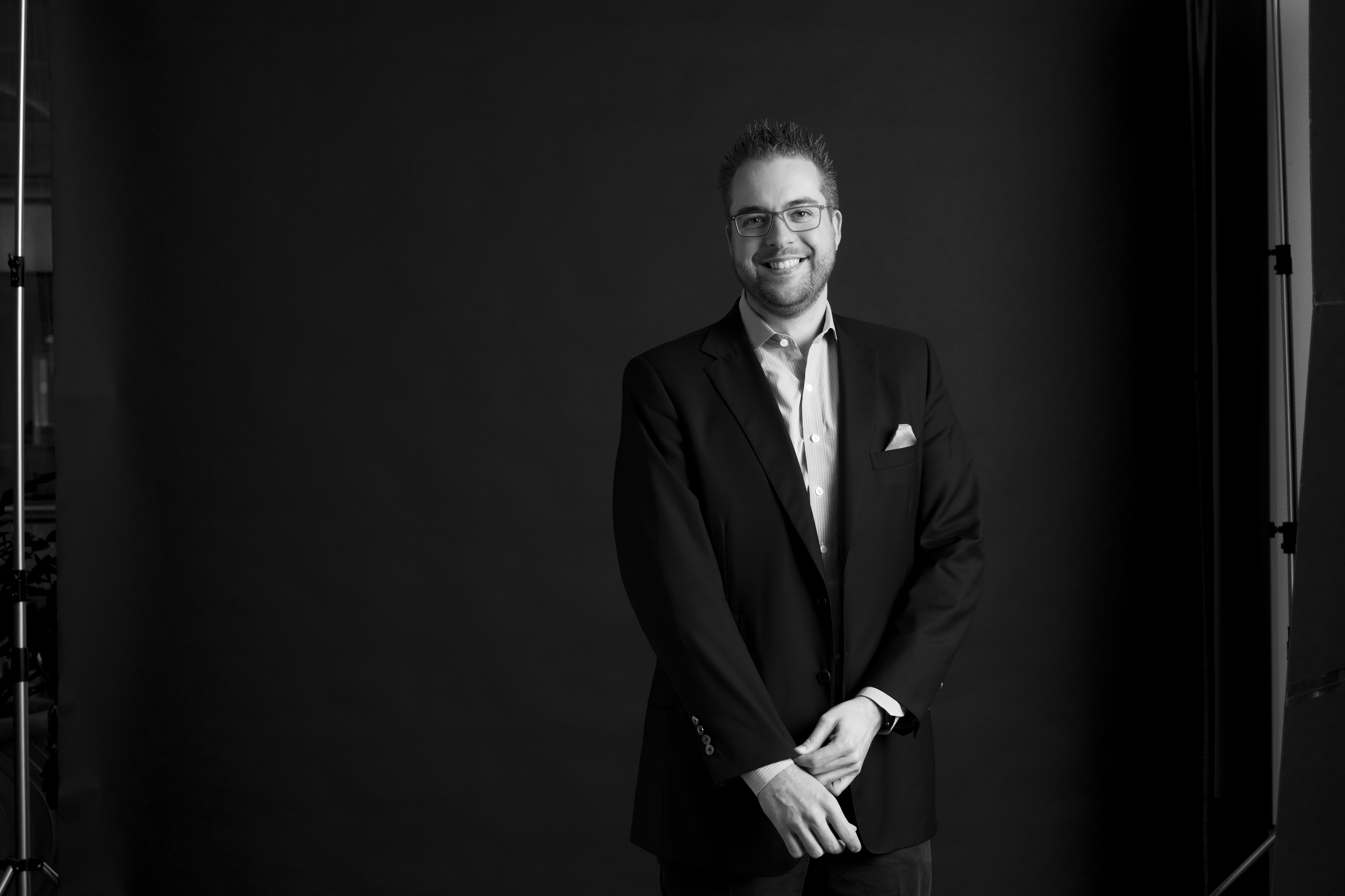A man in a suit and glasses stands in front of a dark backdrop, smiling at the camera.