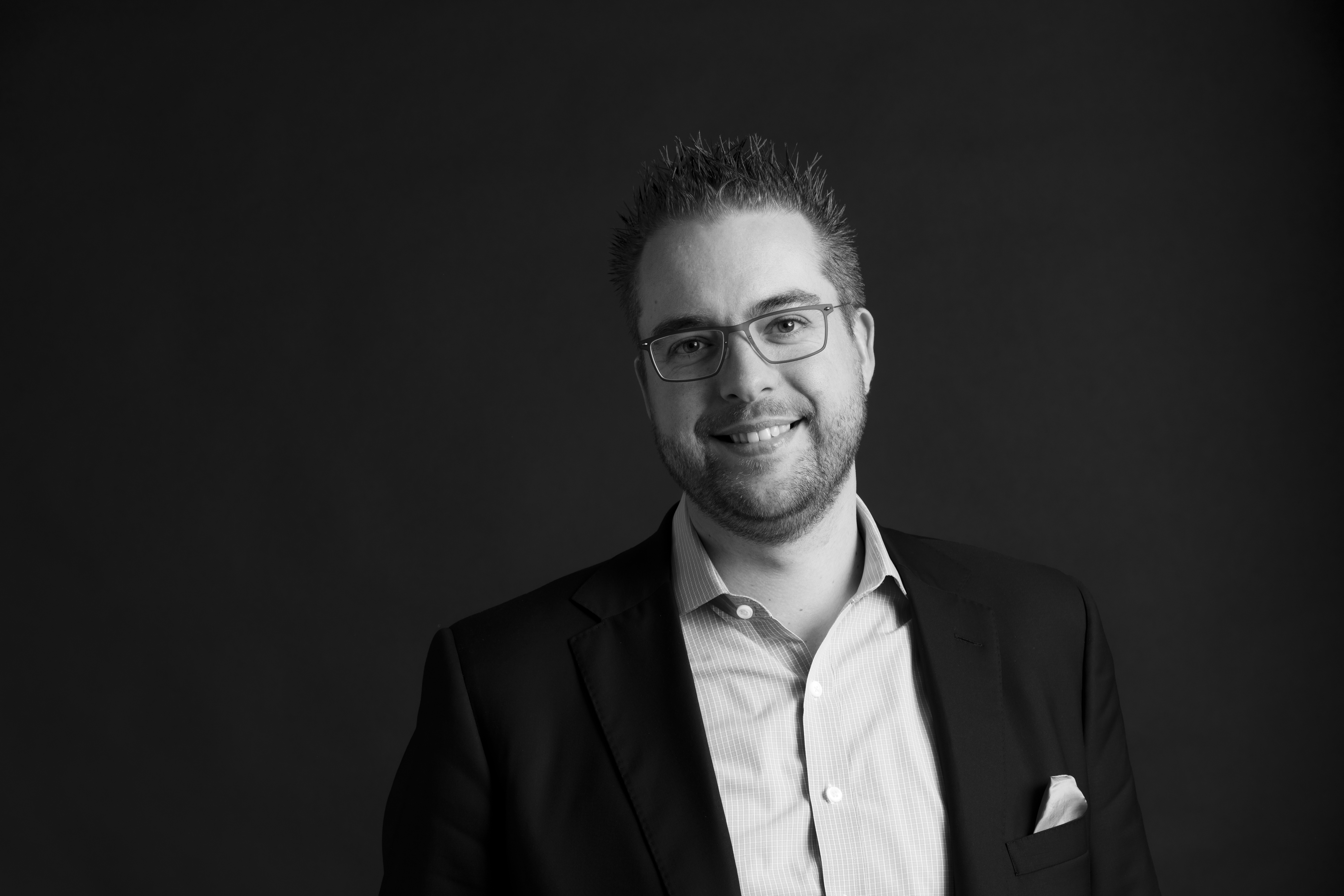 A man wearing glasses and a suit smiles at the camera against a plain dark background.