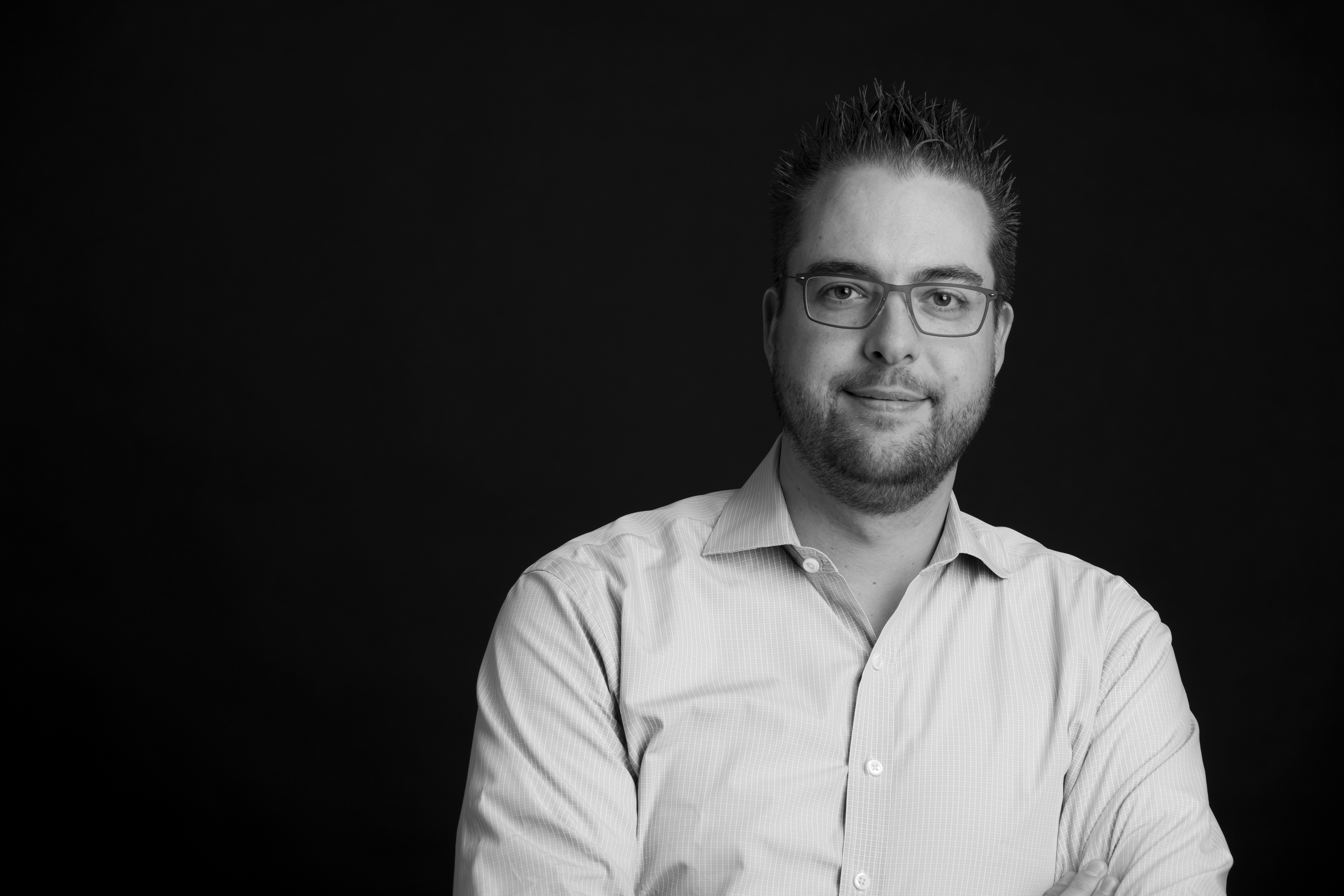 A man wearing glasses and a button-up shirt stands against a dark background, looking directly at the camera.