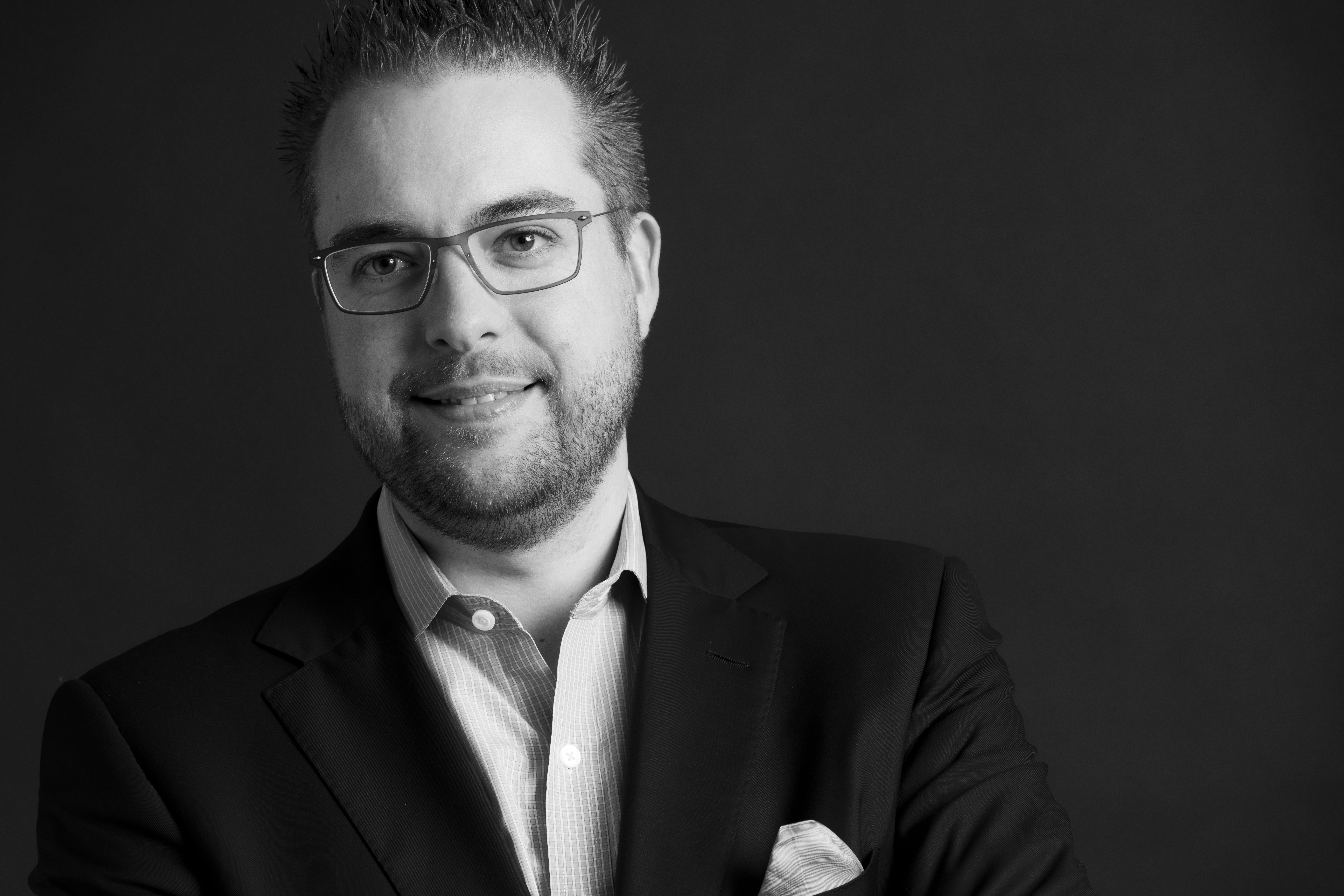 A man wearing glasses and a suit smiles slightly while posing for a professional black-and-white portrait.