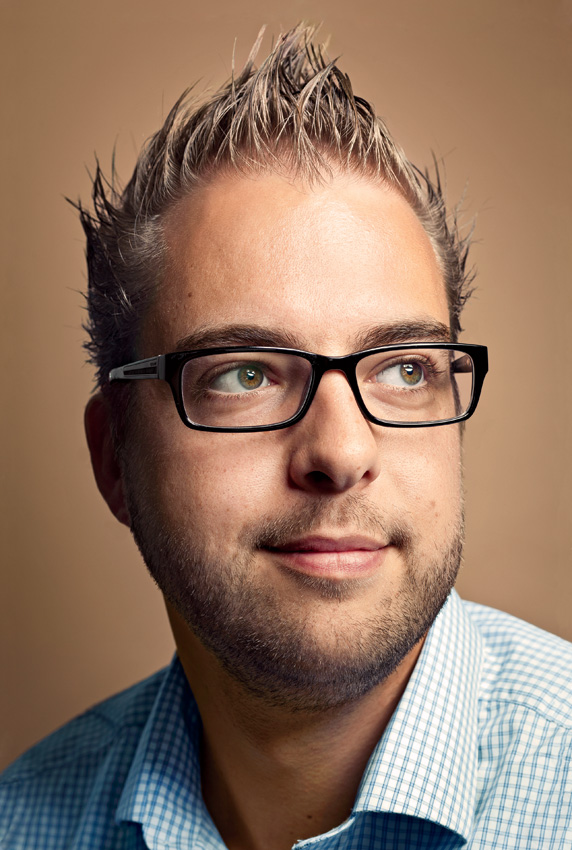 A man with glasses and spiky hair looks slightly to the side, wearing a checkered shirt against a neutral background.