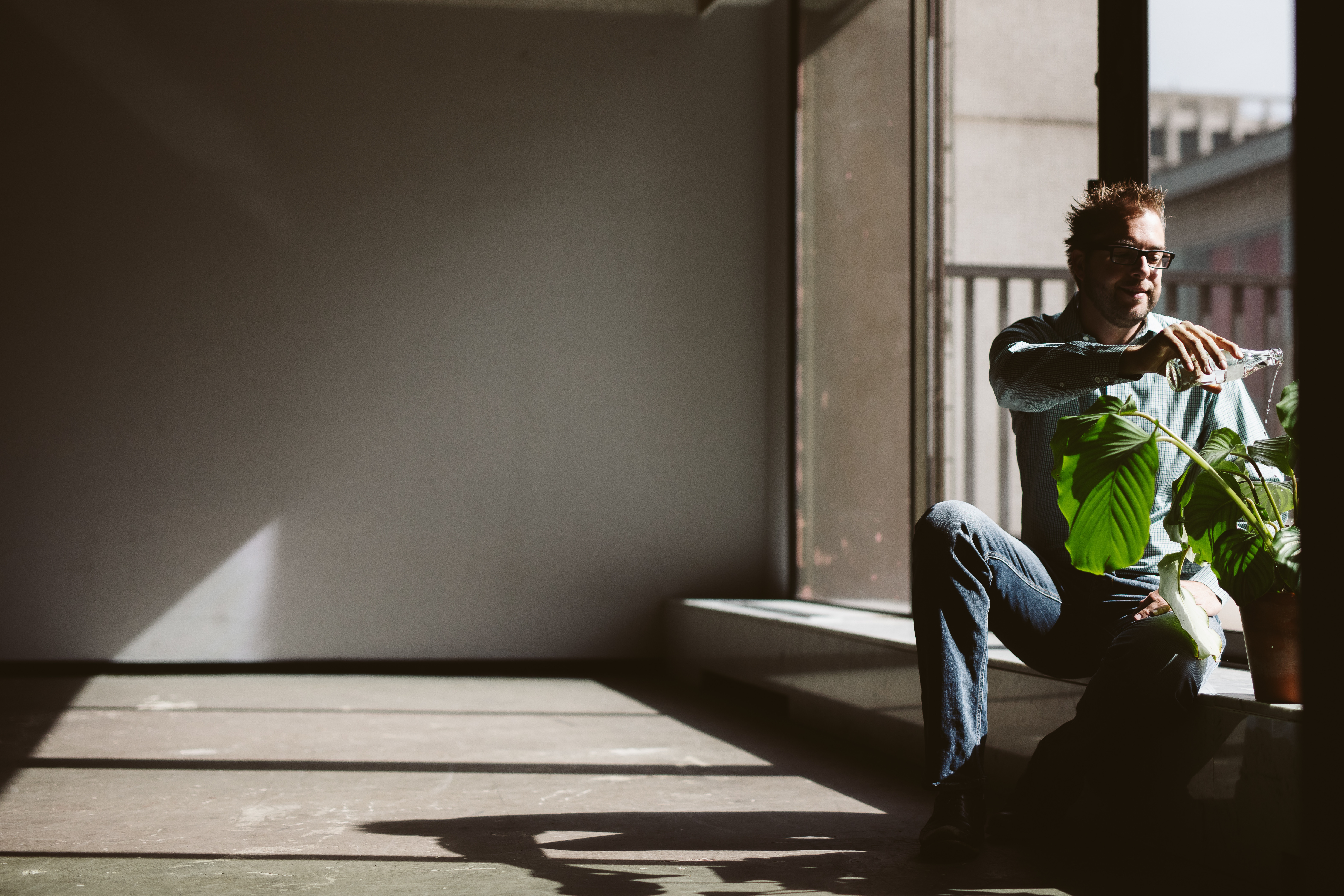 A man wearing glasses sits by a window, pouring water from a bottle onto a potted plant.