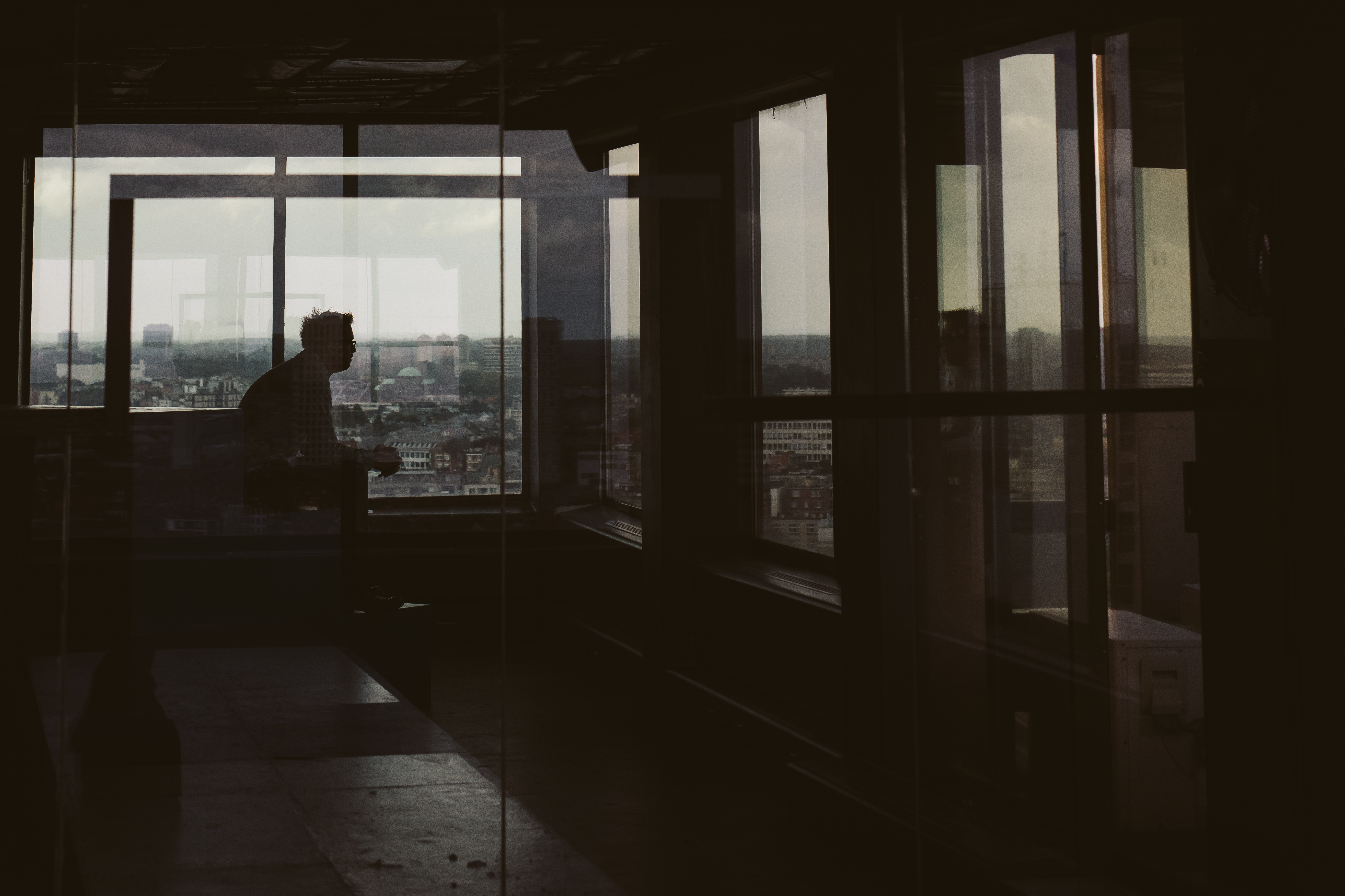 A silhouetted person sits by a large window overlooking a cityscape, with reflections on the glass.