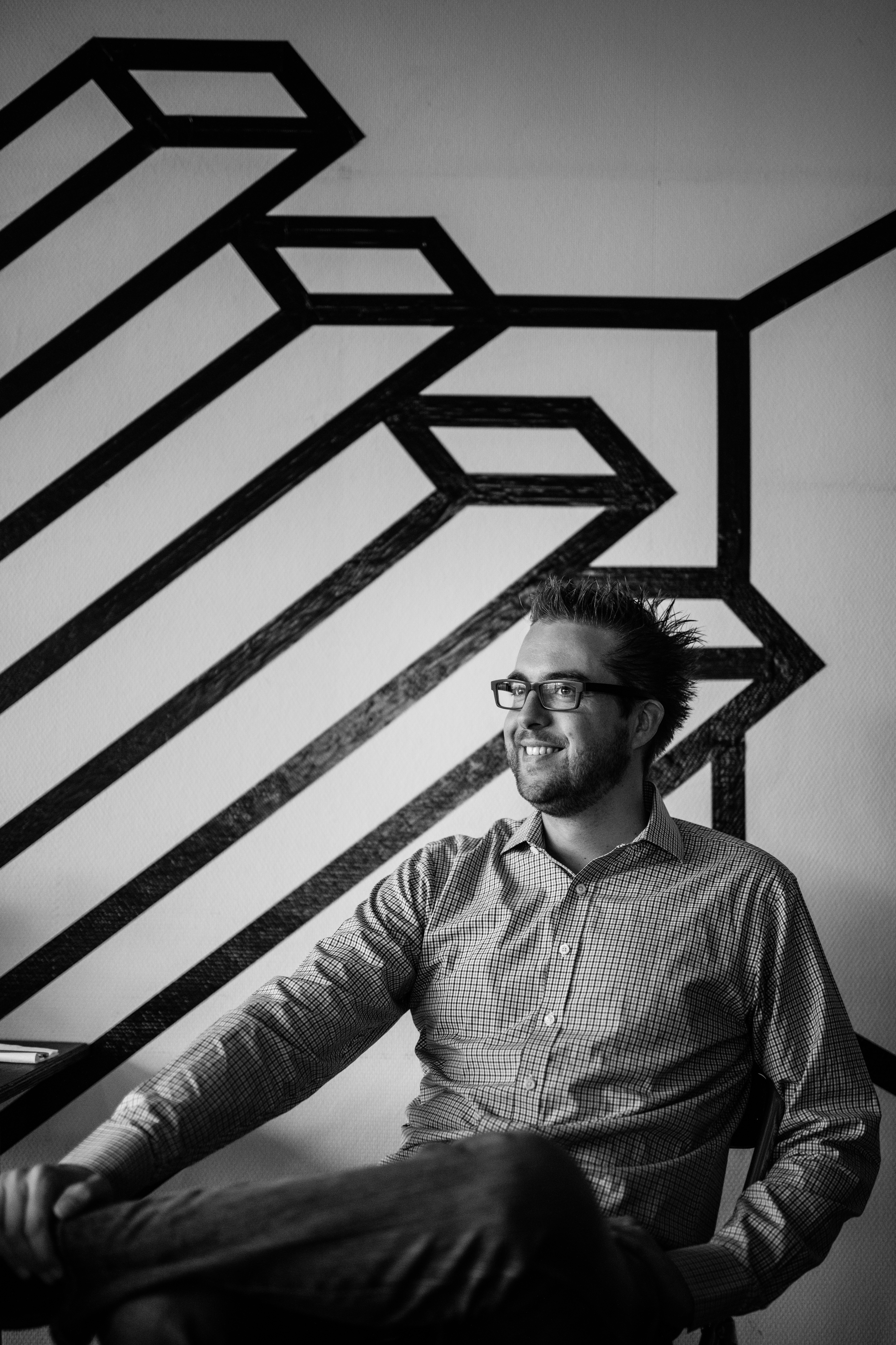 A man wearing glasses and a checkered shirt sits and smiles in front of a geometric wall design.