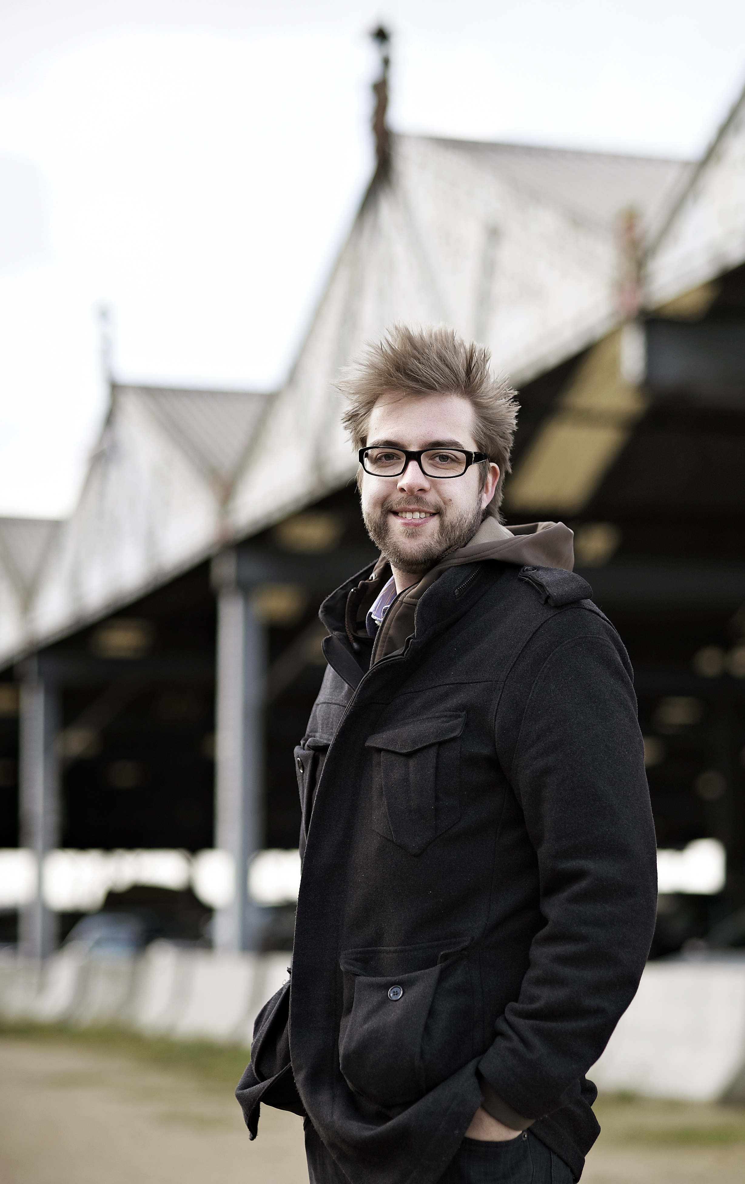 A man with glasses and a black coat stands outdoors, smiling at the camera.