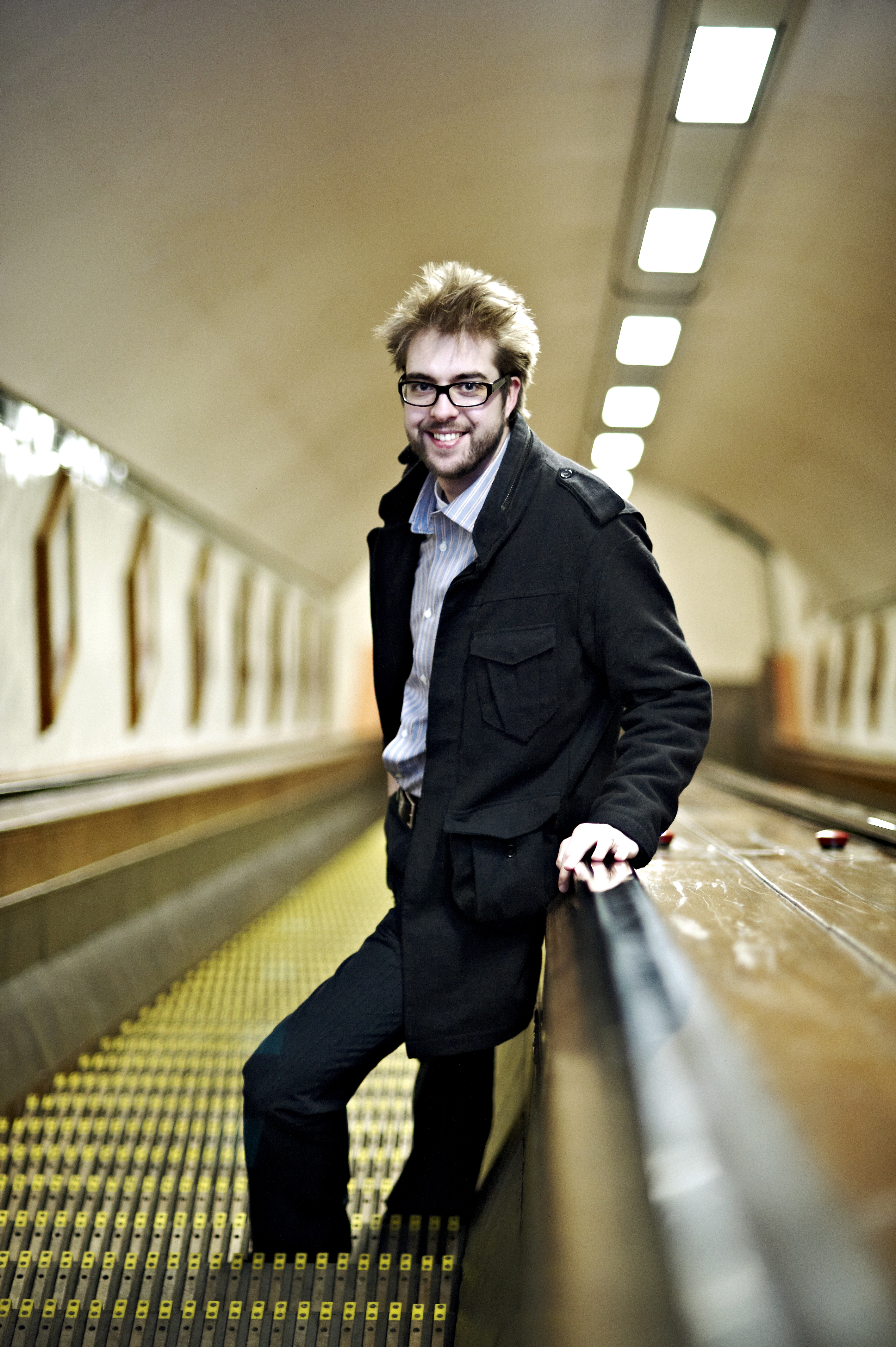 A man with glasses and a black coat smiles while leaning on a handrail in a tunnel with escalators.
