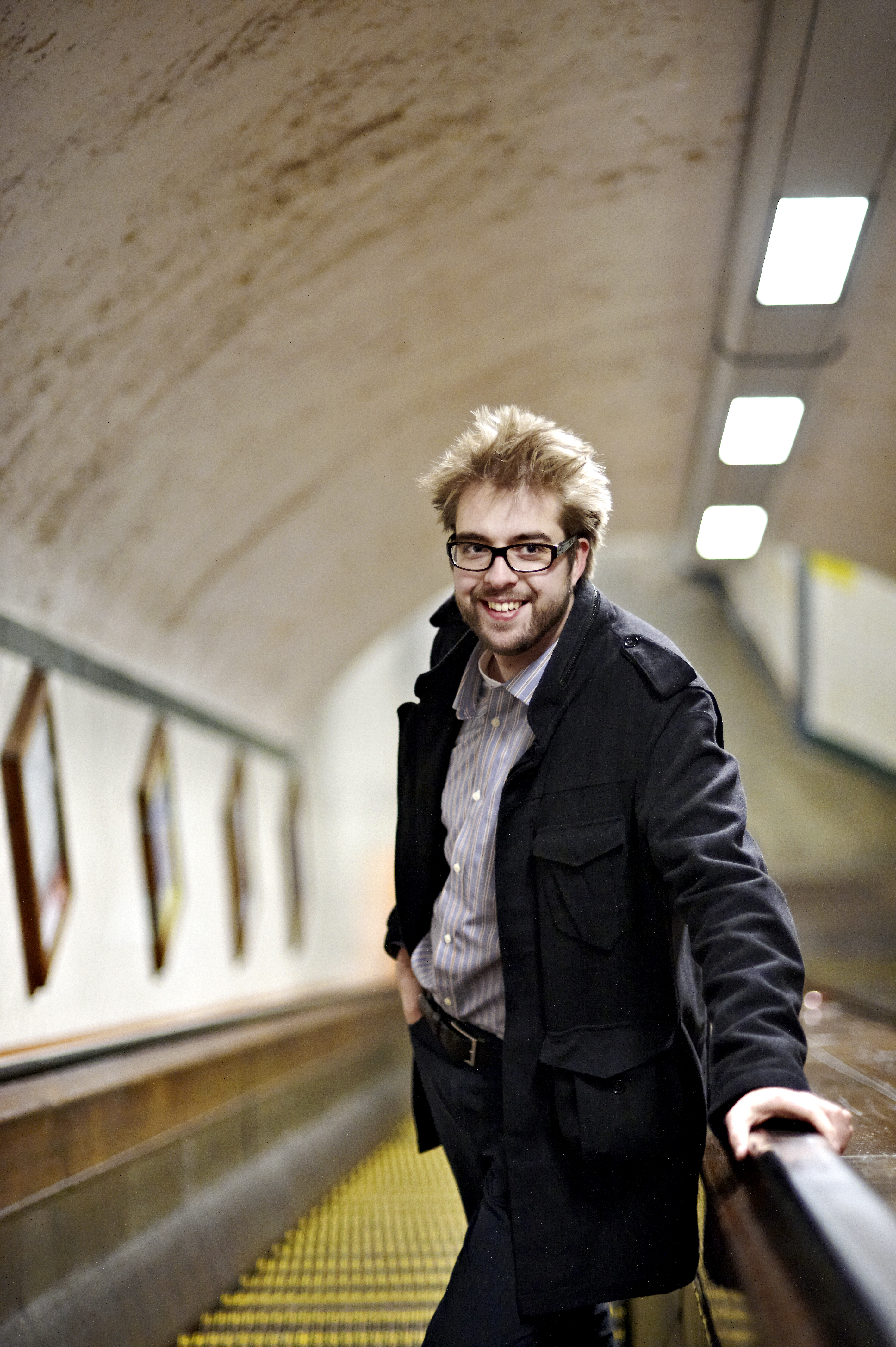 A man with glasses and a black coat smiles while leaning on an escalator handrail in a tunnel.