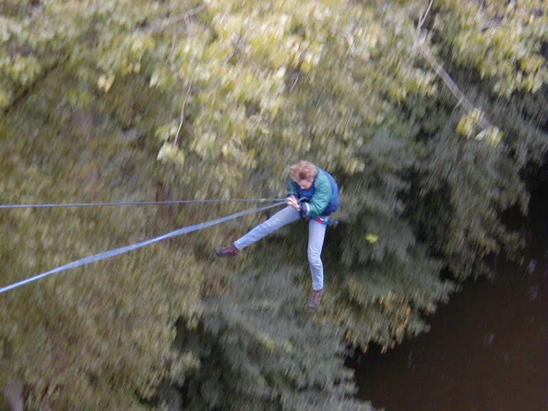 A person in a harness swings from a rope over a river, holding on tightly with both hands.