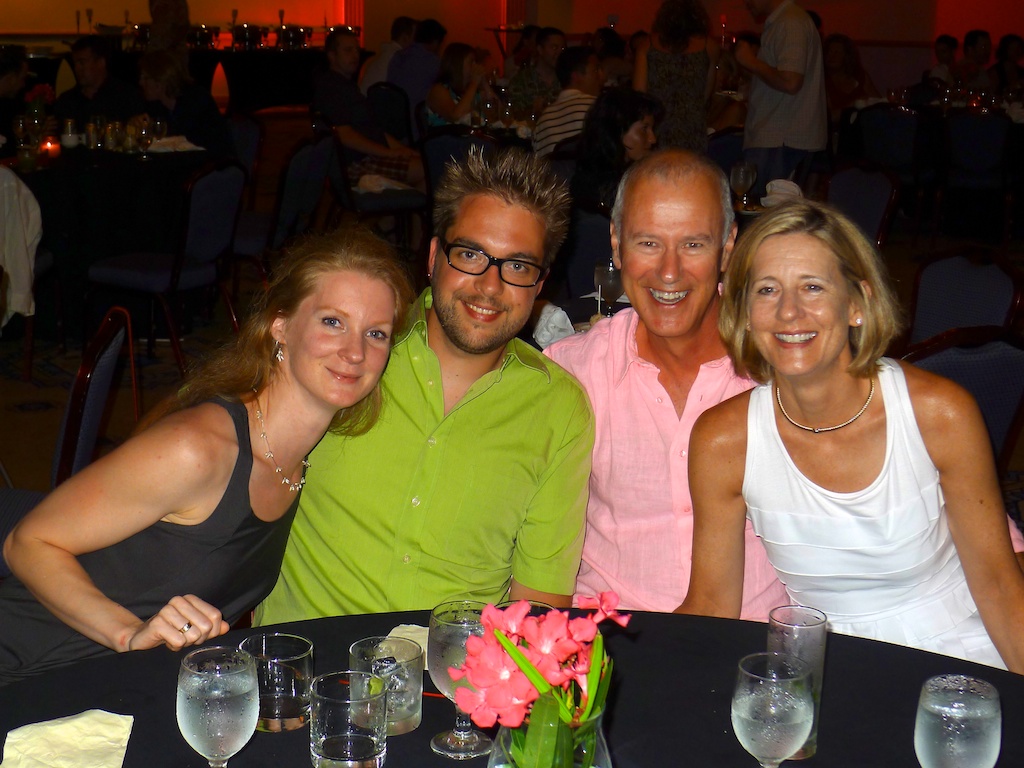 Four people sitting at a table, smiling at the camera, with drinks and flowers in front of them.