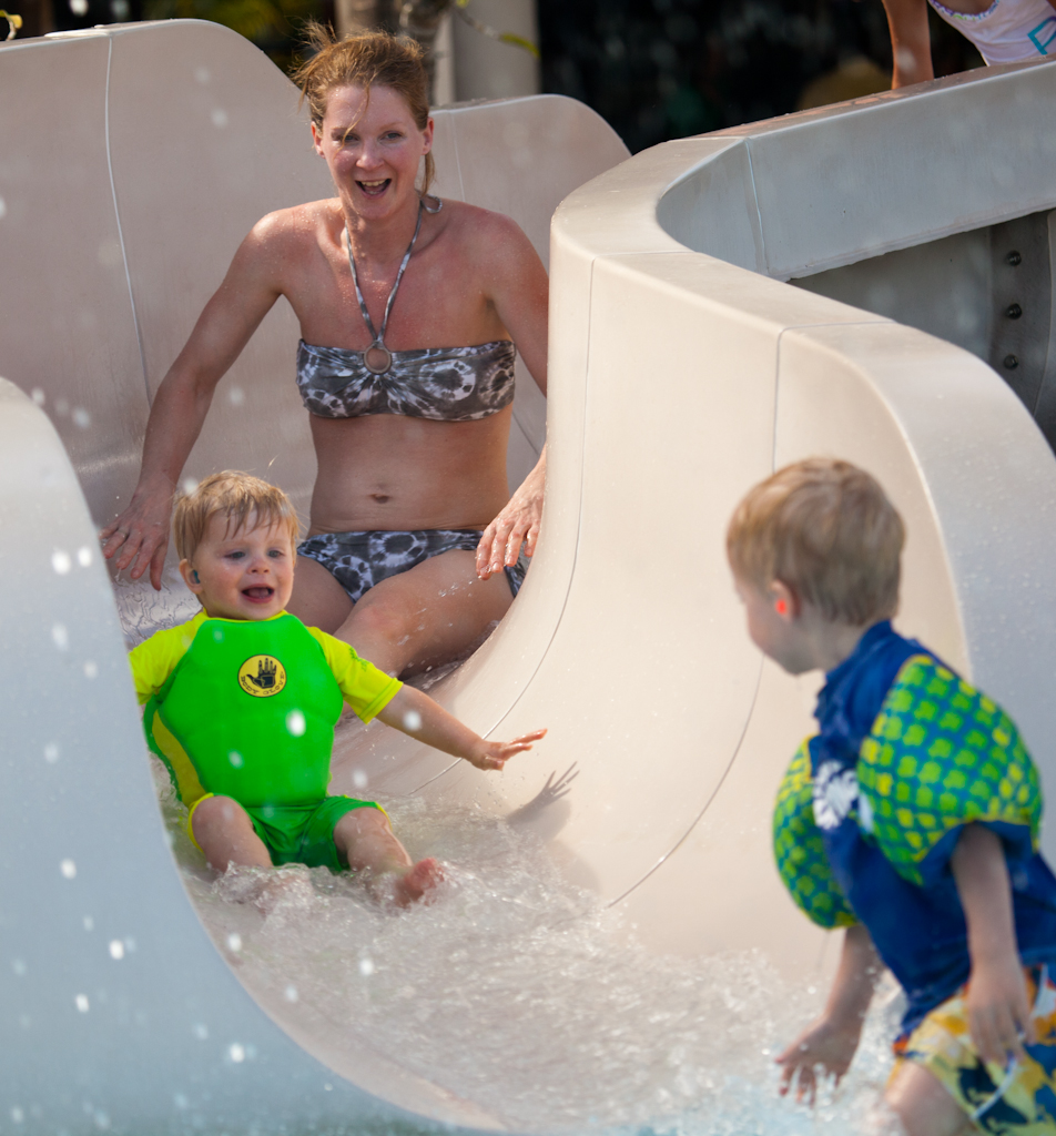 A smiling child in a green swimsuit slides down a water slide, followed by an adult, while another child watches.