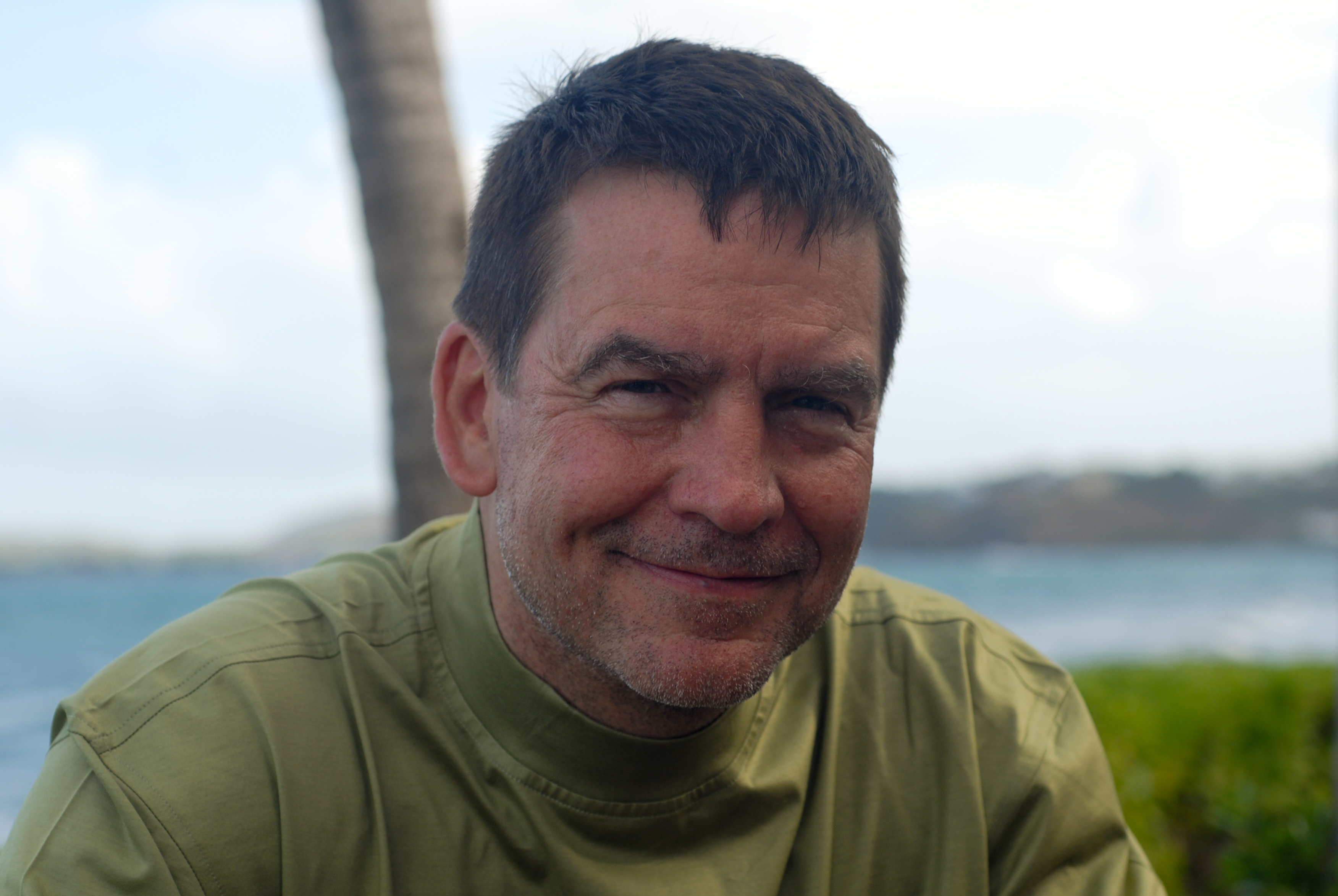 A man in a green shirt smiles at the camera with a blurred ocean and palm tree in the background.