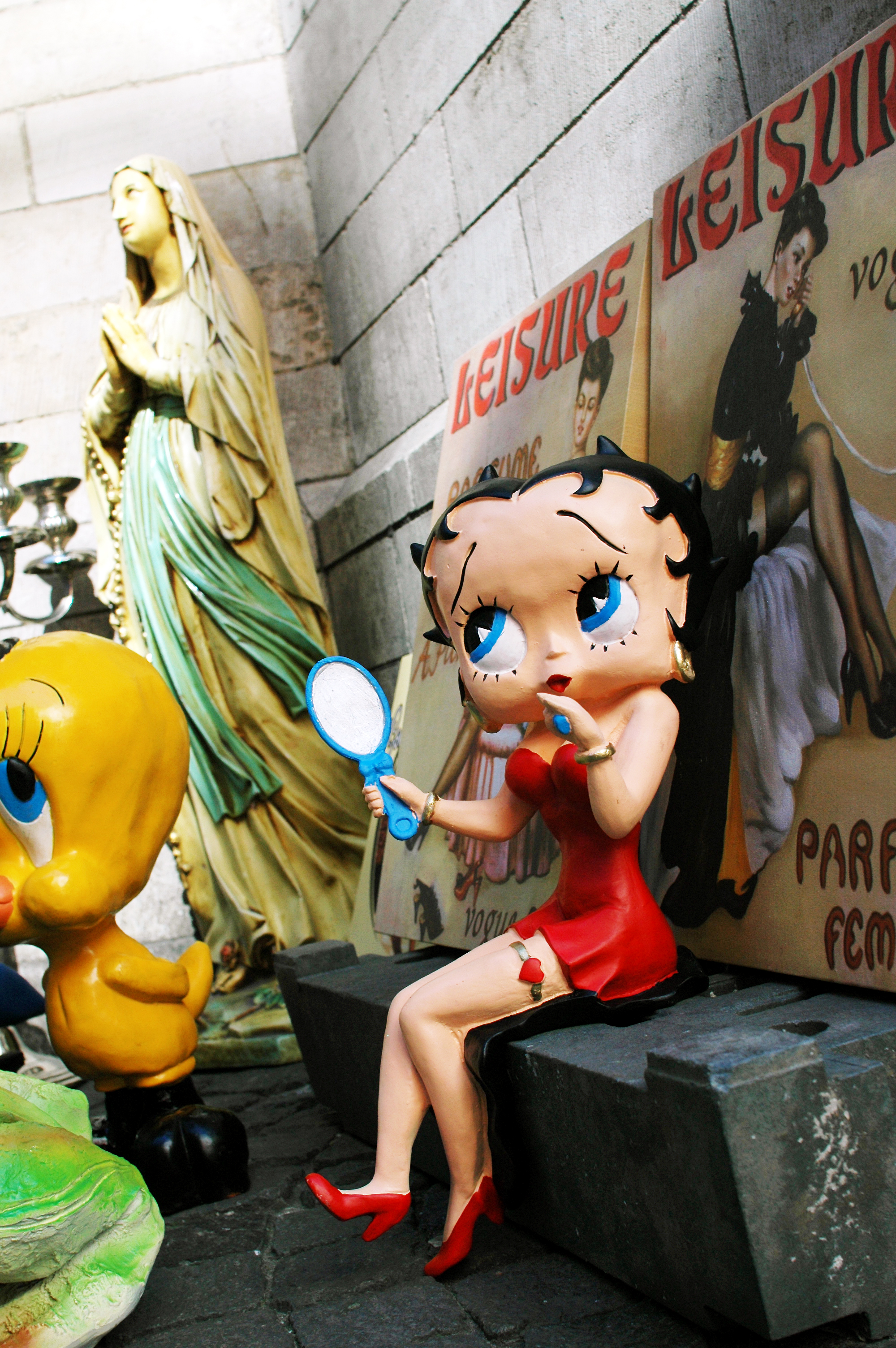 A statue of Betty Boop in a red dress sits on a bench, holding a mirror and applying lipstick.