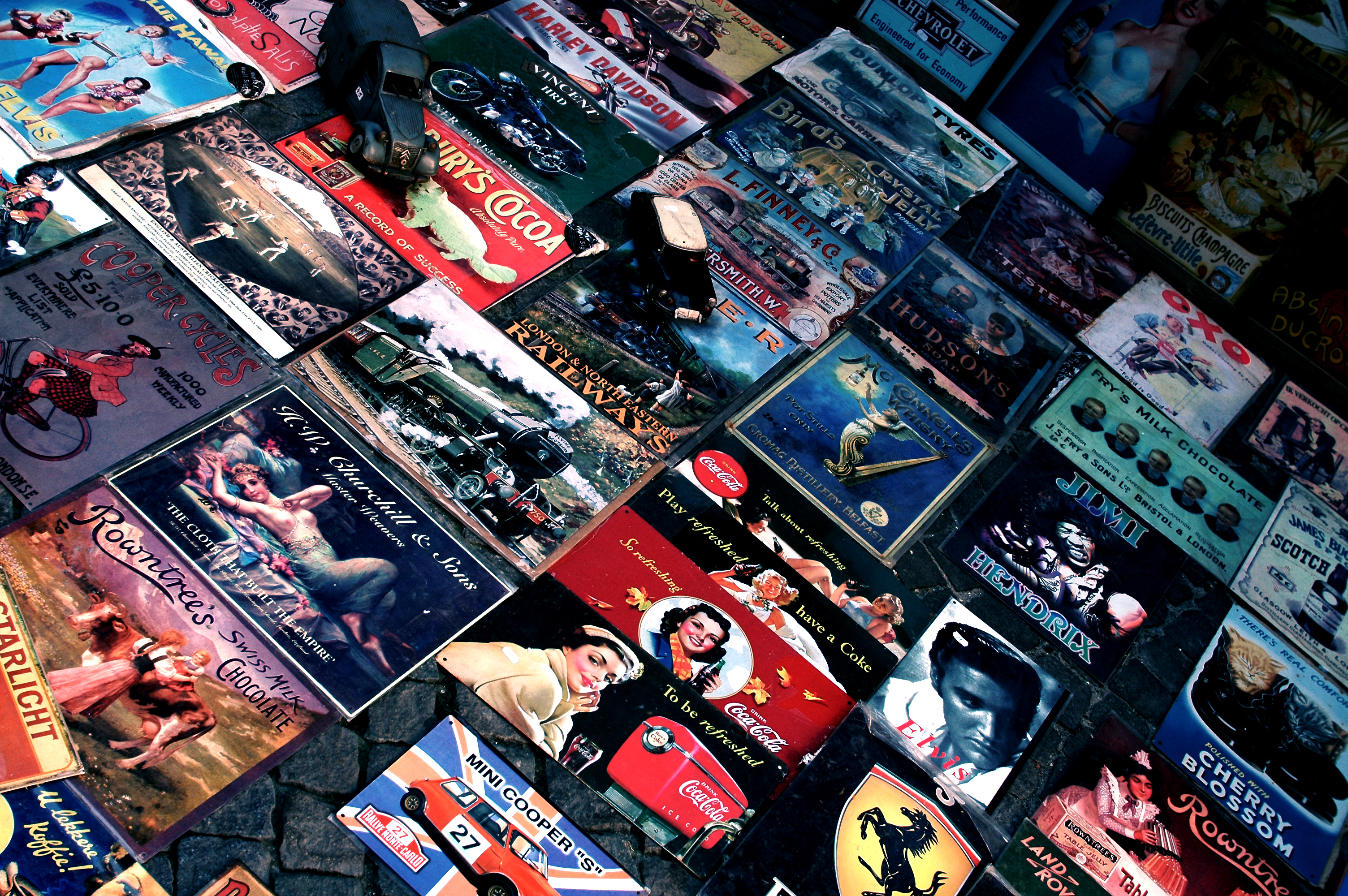 A collection of vintage metal signs with various advertisements and images displayed on a cobblestone surface.
