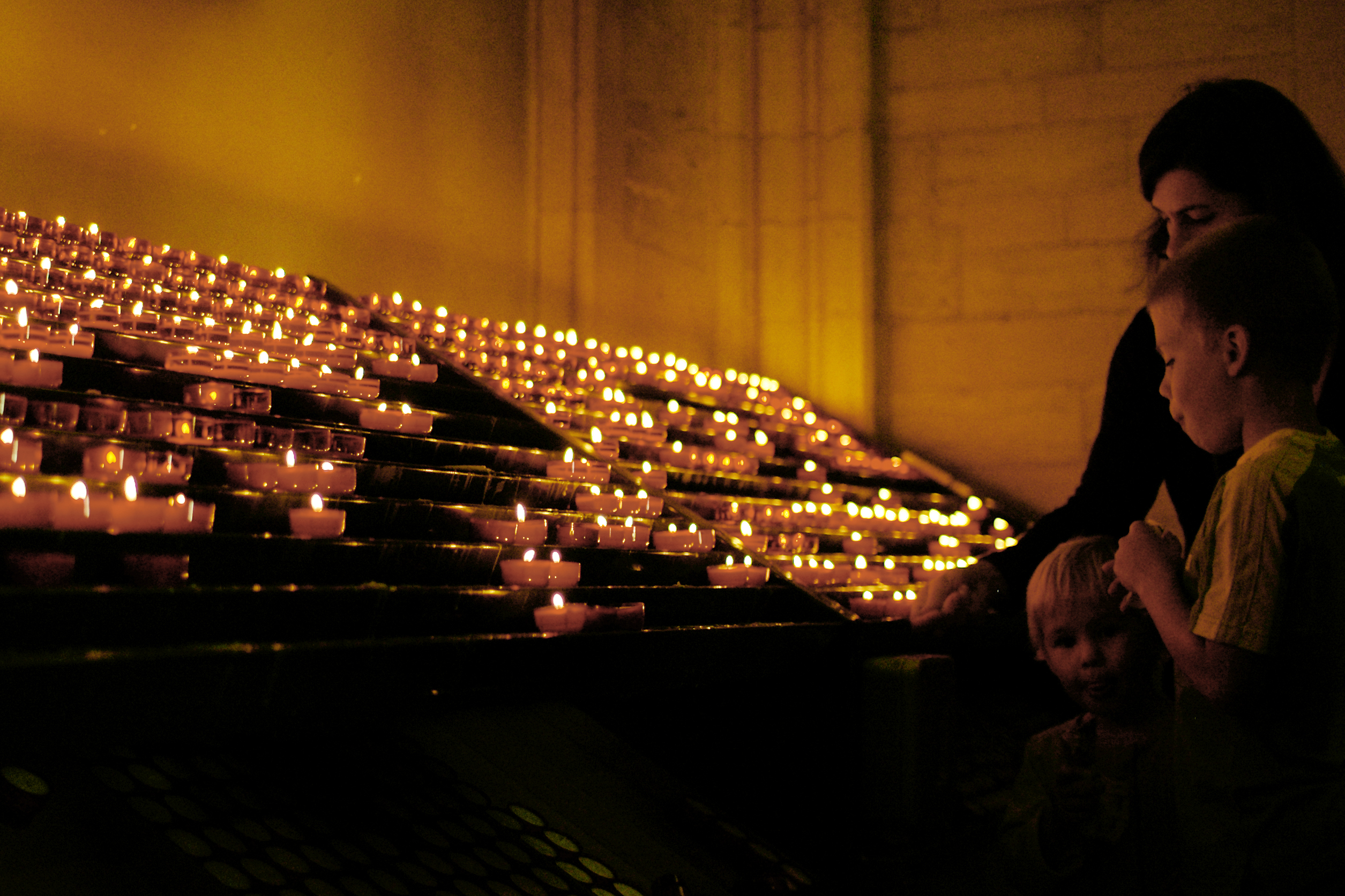 A woman and two children stand near rows of lit candles, appearing to observe or participate in a ritual.