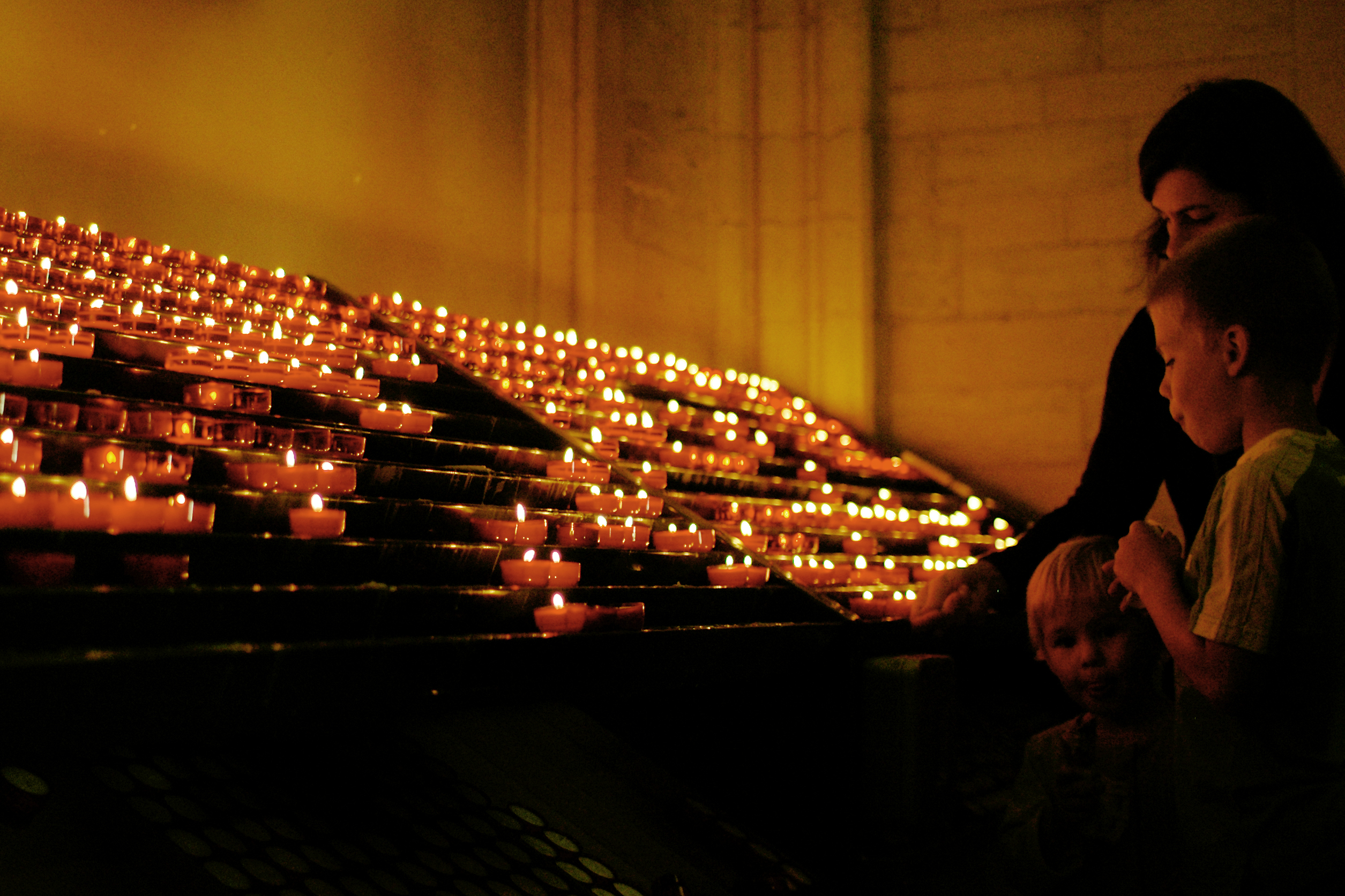 A woman and two children stand near a display of lit candles in a dimly lit space.