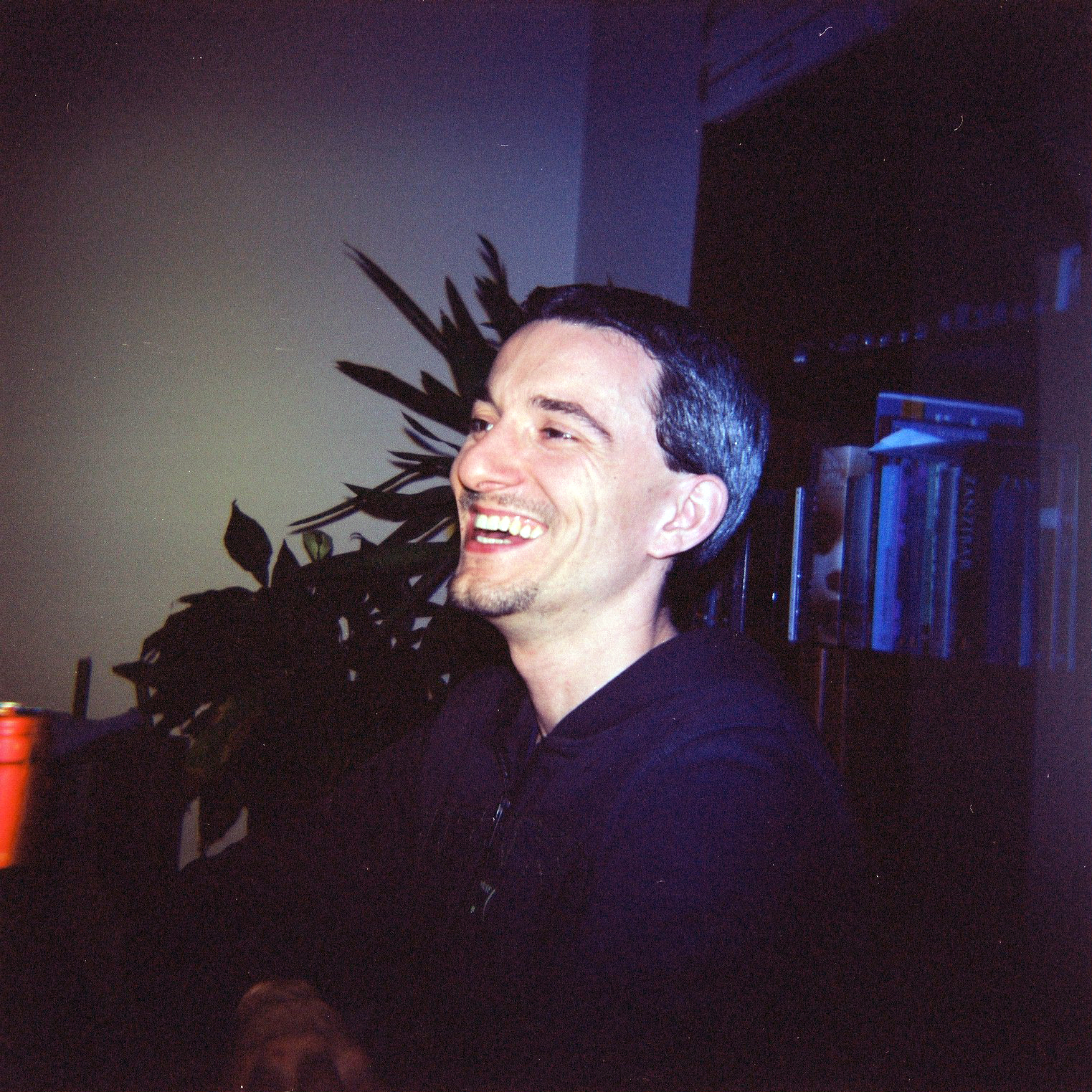 A man with short dark hair and a beard is laughing while sitting indoors.