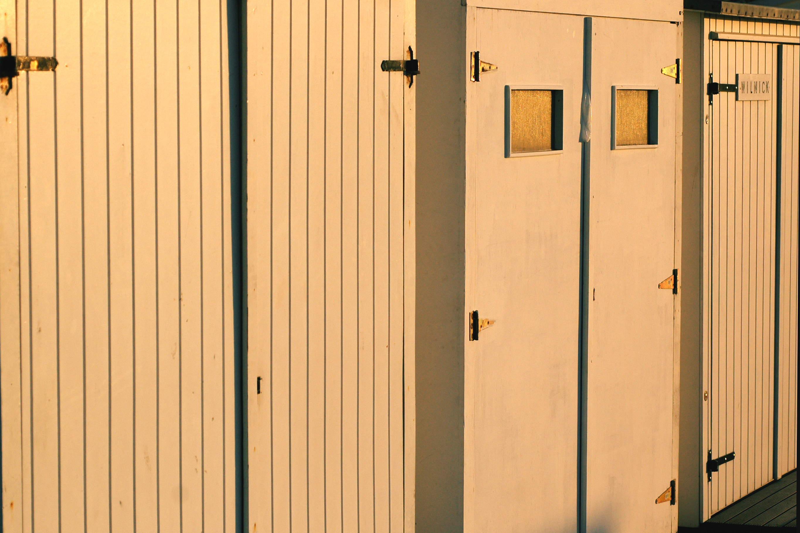 A row of wooden cabin doors with metal hinges, some featuring small windows, illuminated by warm sunlight.