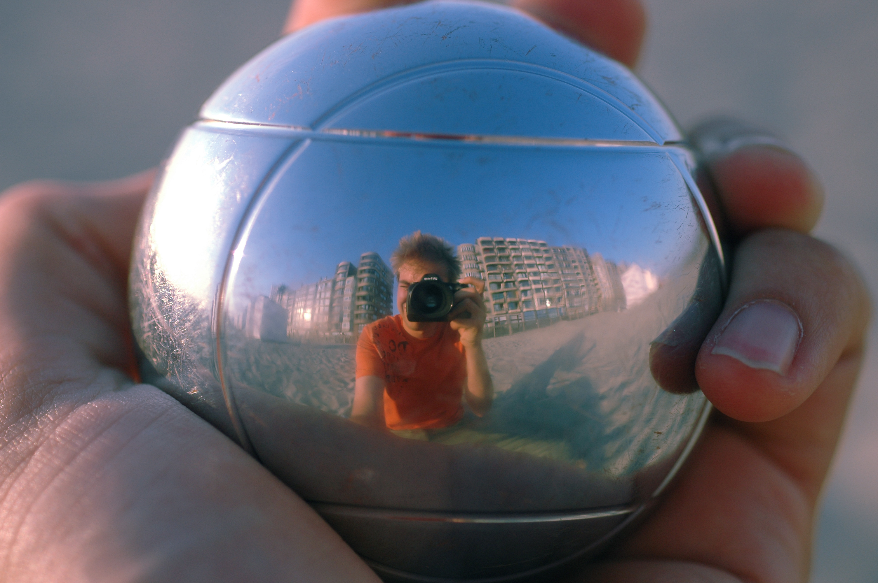 A hand holds a reflective metal ball, showing a distorted reflection of a person taking a photo with a camera.